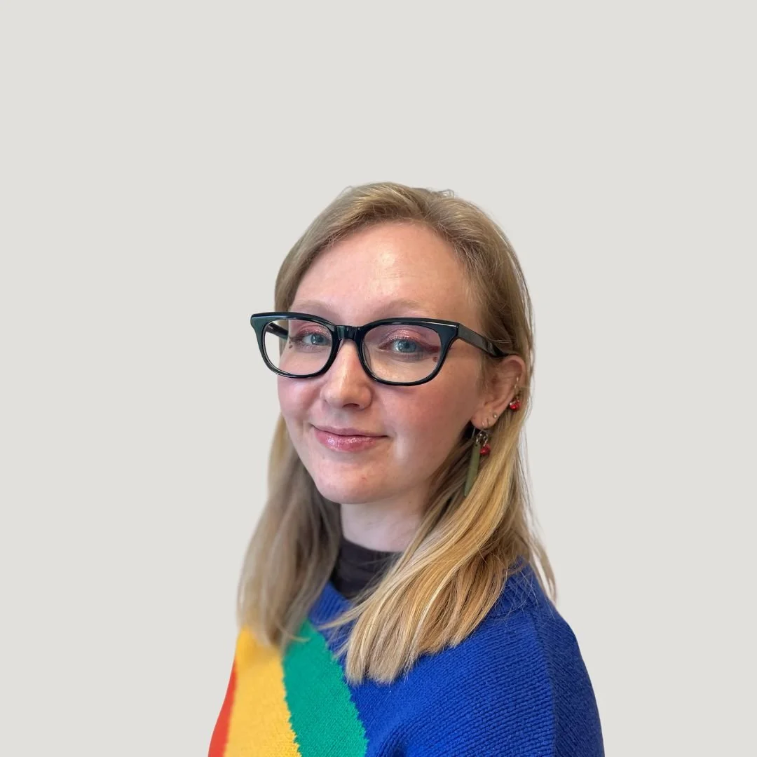 A woman with blonde hair, black glasses, and earrings, wearing a rainbow-colored sweater, smiling against a plain white background.
