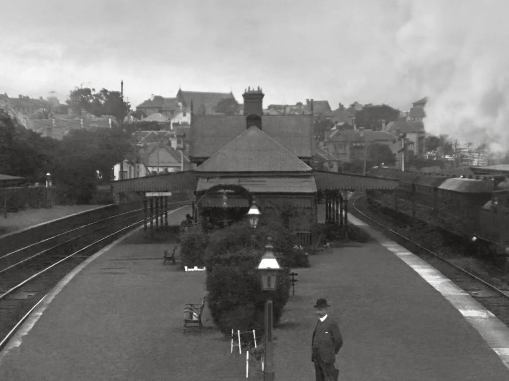 Black and white photo of train station platform with a man in early 20th-century attire, benches, trees, and a train on the right side, with historical buildings in the background.