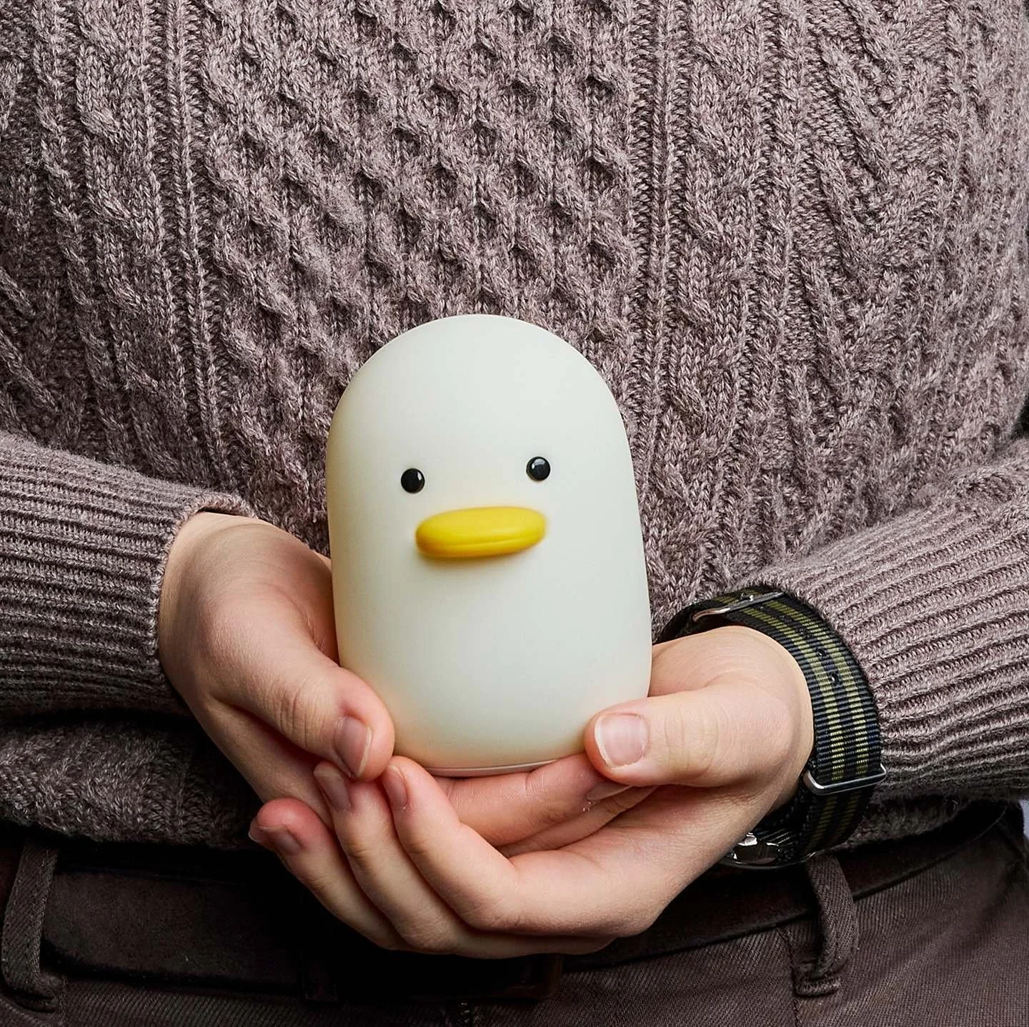 Person wearing a brown knitted sweater and a wristwatch holding a small cream-colored duck-shaped object with black eyes and a yellow beak.