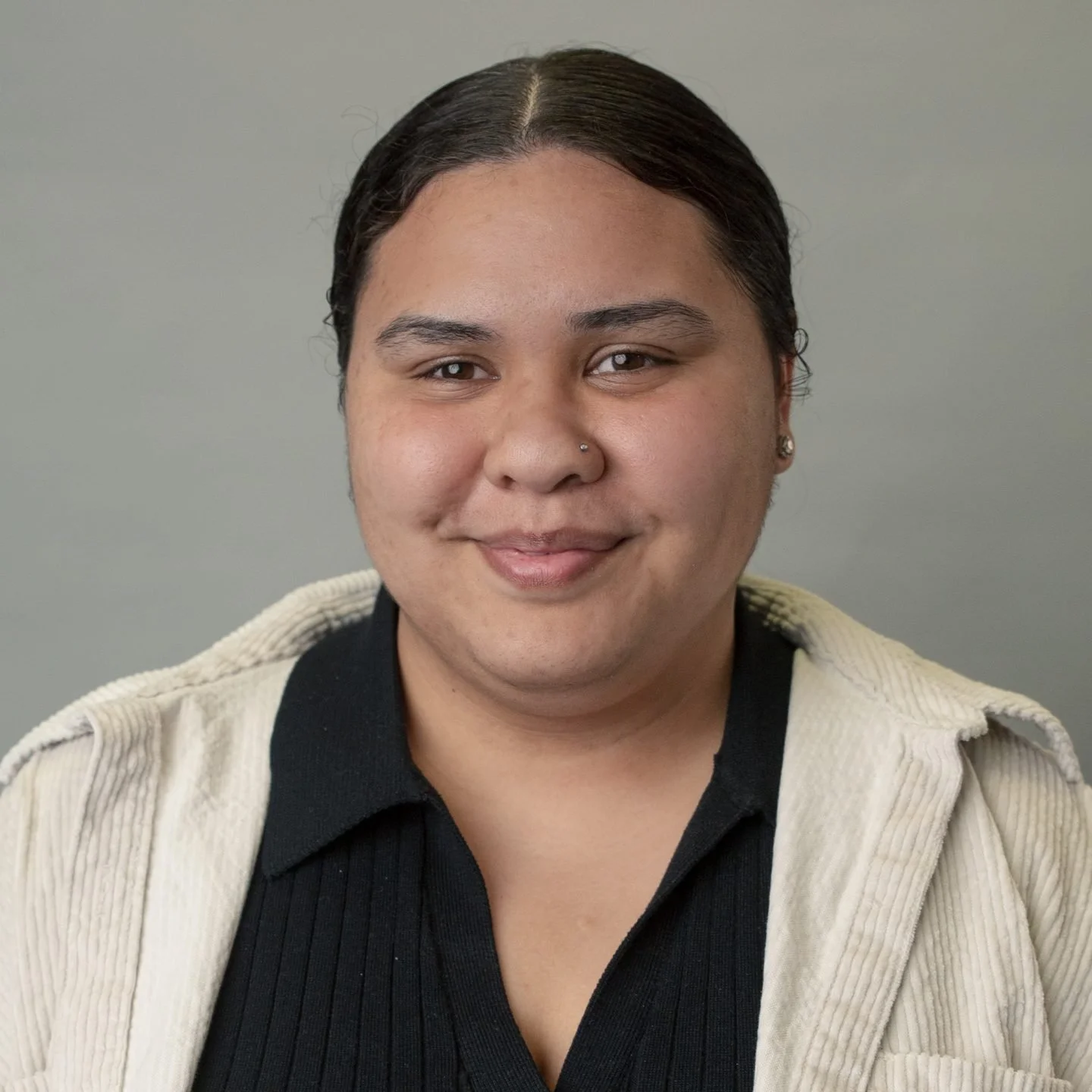 A person with dark hair, a nose piercing, and earrings, smiling with closed lips, wearing a black shirt and a beige jacket against a gray background.