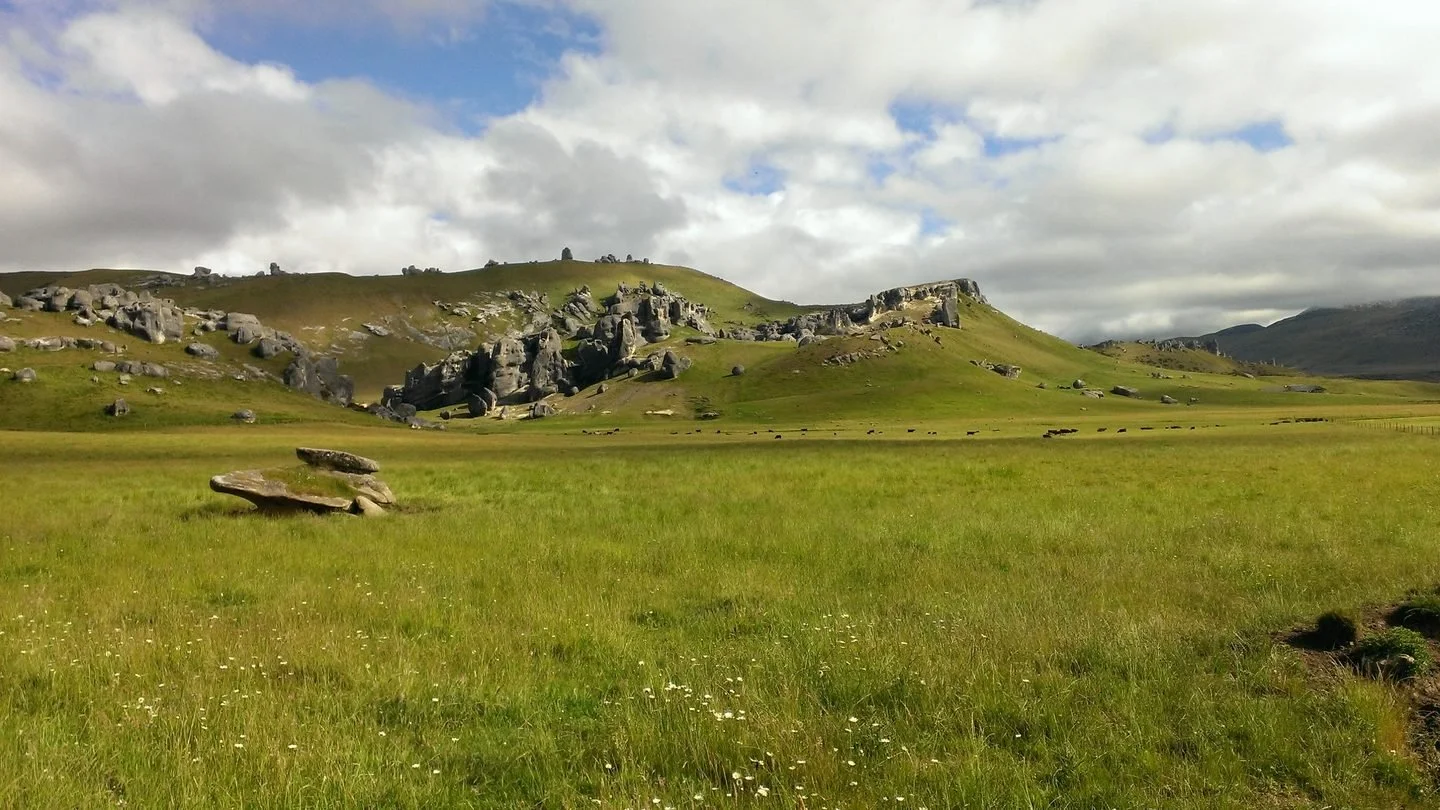 A vast green field with rocks scattered, leading to rocky hills under a partly cloudy sky.