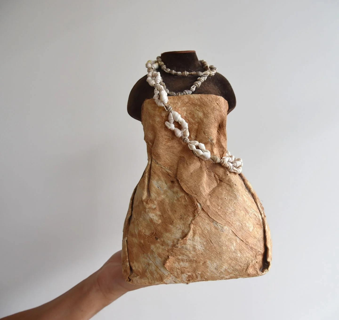 A jewelry mannequin adorned with pearl necklaces, draped over a textured beige cloth covering a dark mannequin bust, held by a hand against a plain white background.