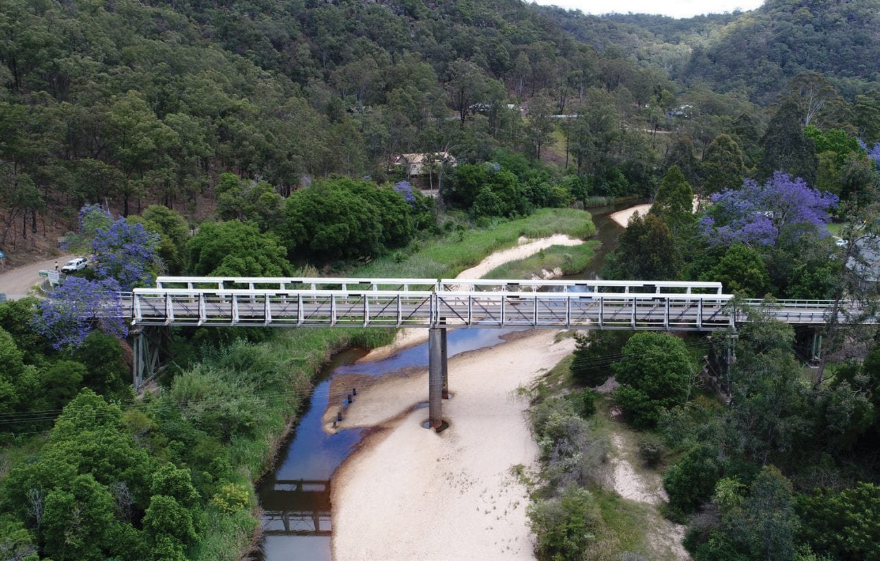 A river flowing under a metal bridge in a green, hilly landscape with trees and purple flowering trees.