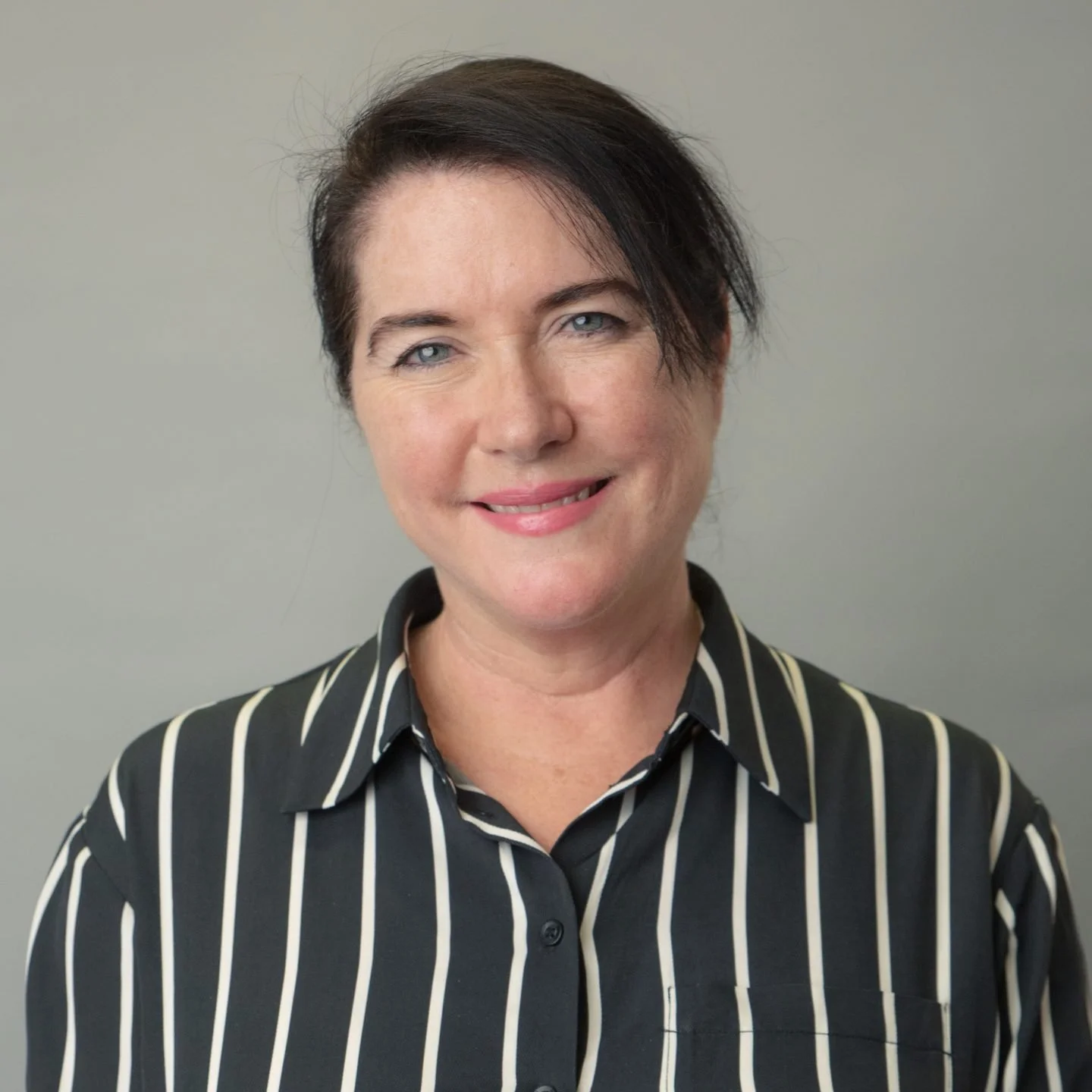 A woman with short dark hair and blue eyes wearing a black and white striped button-up shirt, smiling against a plain gray background.