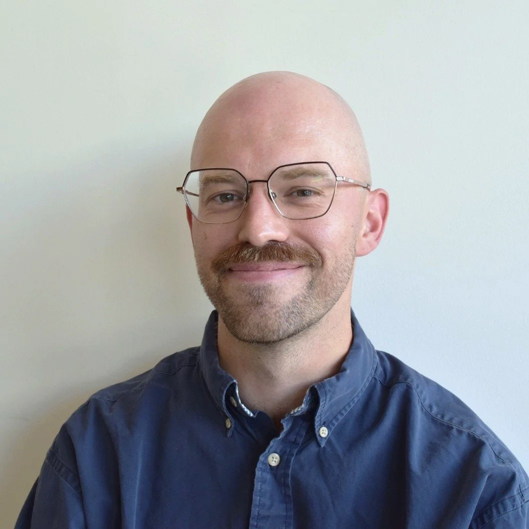 A man with a bald head, glasses, and a beard, smiling and wearing a blue button-up shirt, standing against a plain white wall.