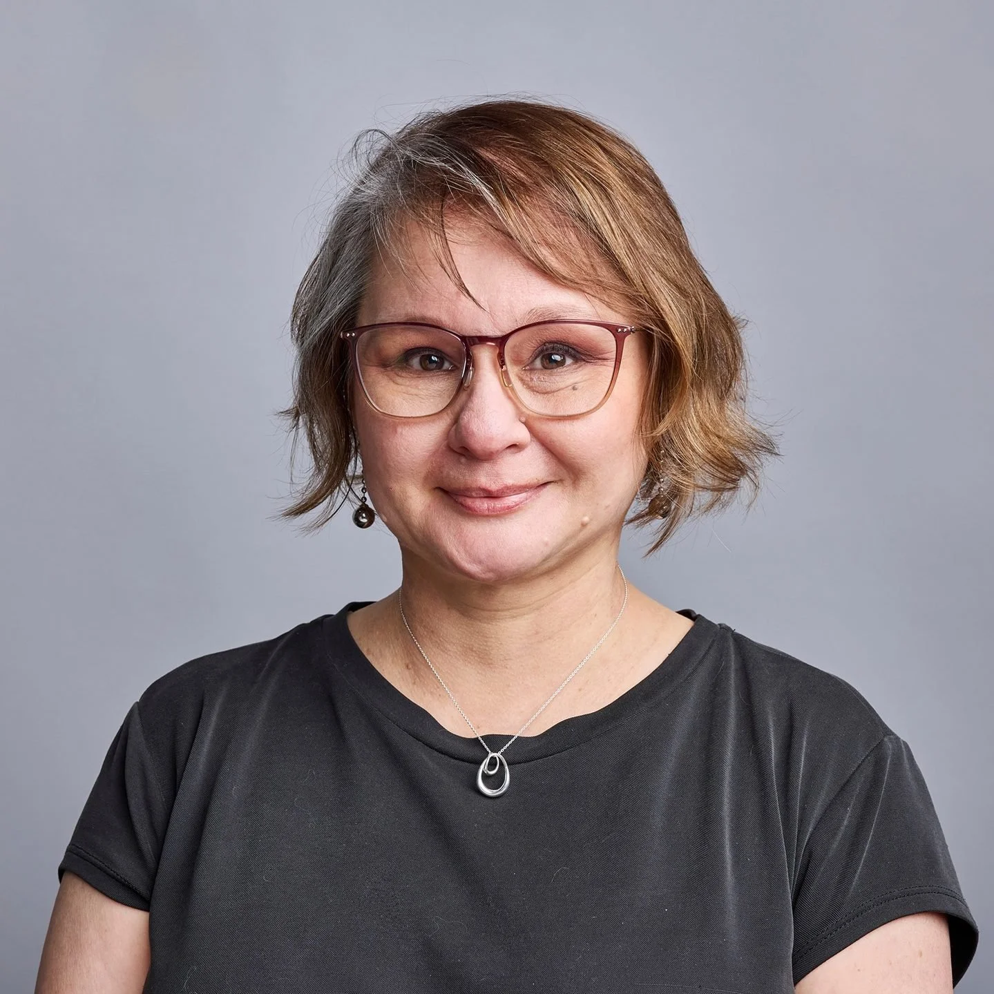 A middle-aged woman with short, wavy, light brown hair wearing glasses, a black t-shirt, and a silver necklace, smiling at the camera against a plain gray background.