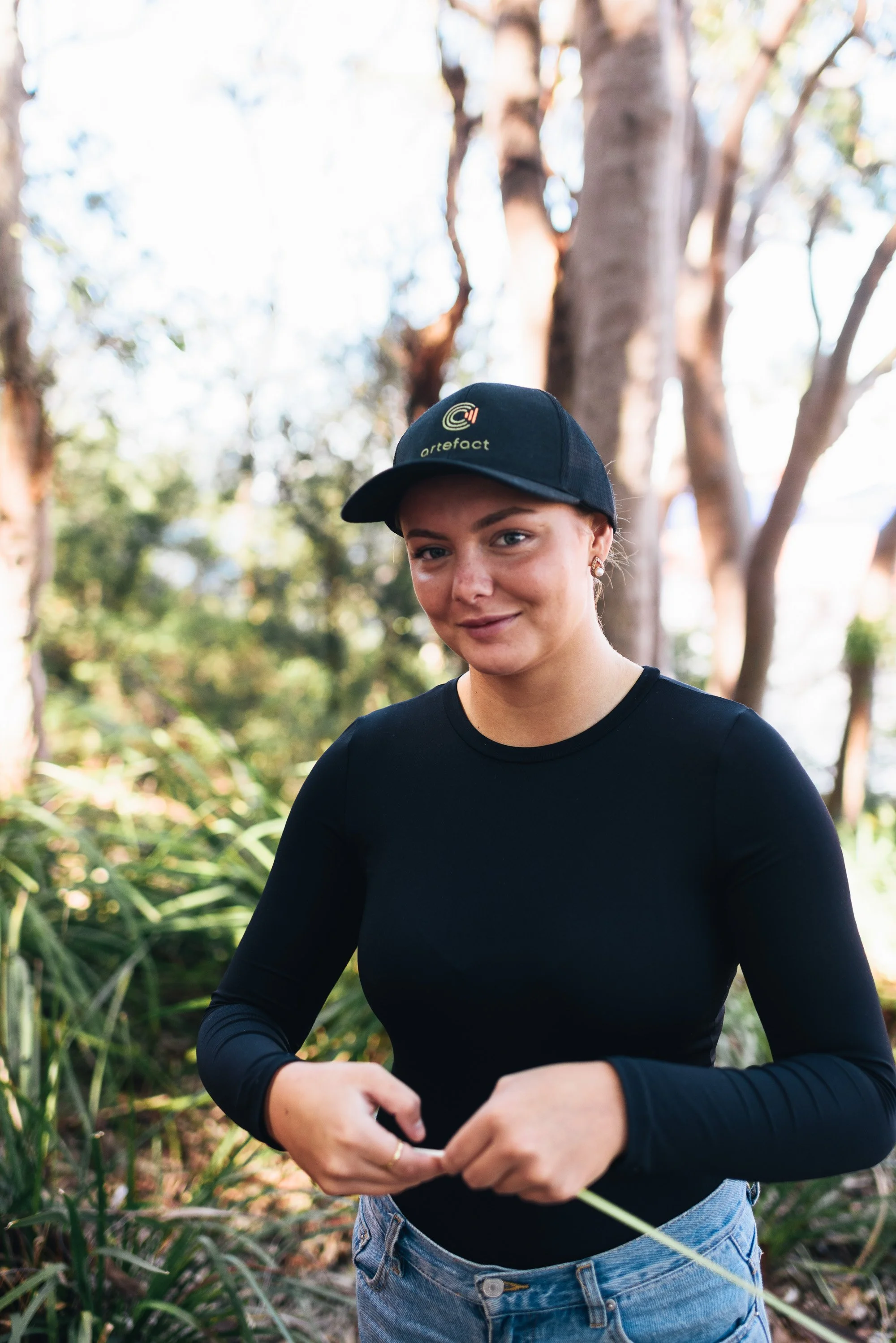 A young woman with fair skin and blonde hair, wearing a black cap with a logo and the word 'artefact', a black long-sleeve shirt, and light blue jeans, stands outdoors in a natural setting with trees and greenery in the background. She is smiling slightly and looking at the camera.