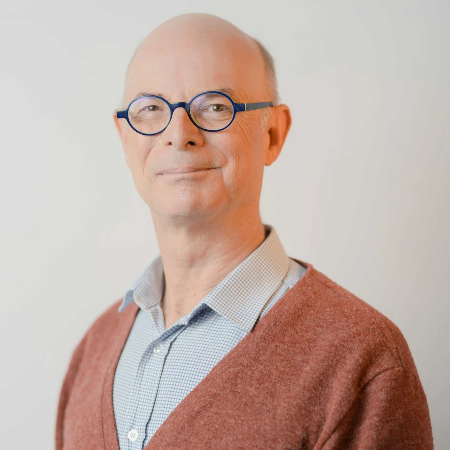 A middle-aged man with glasses and a bald head, smiling slightly, wearing a checkered shirt and a rust-colored sweater.
