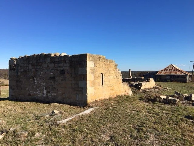 Old stone building ruins in a grassy area under a clear blue sky with other structures in the background.