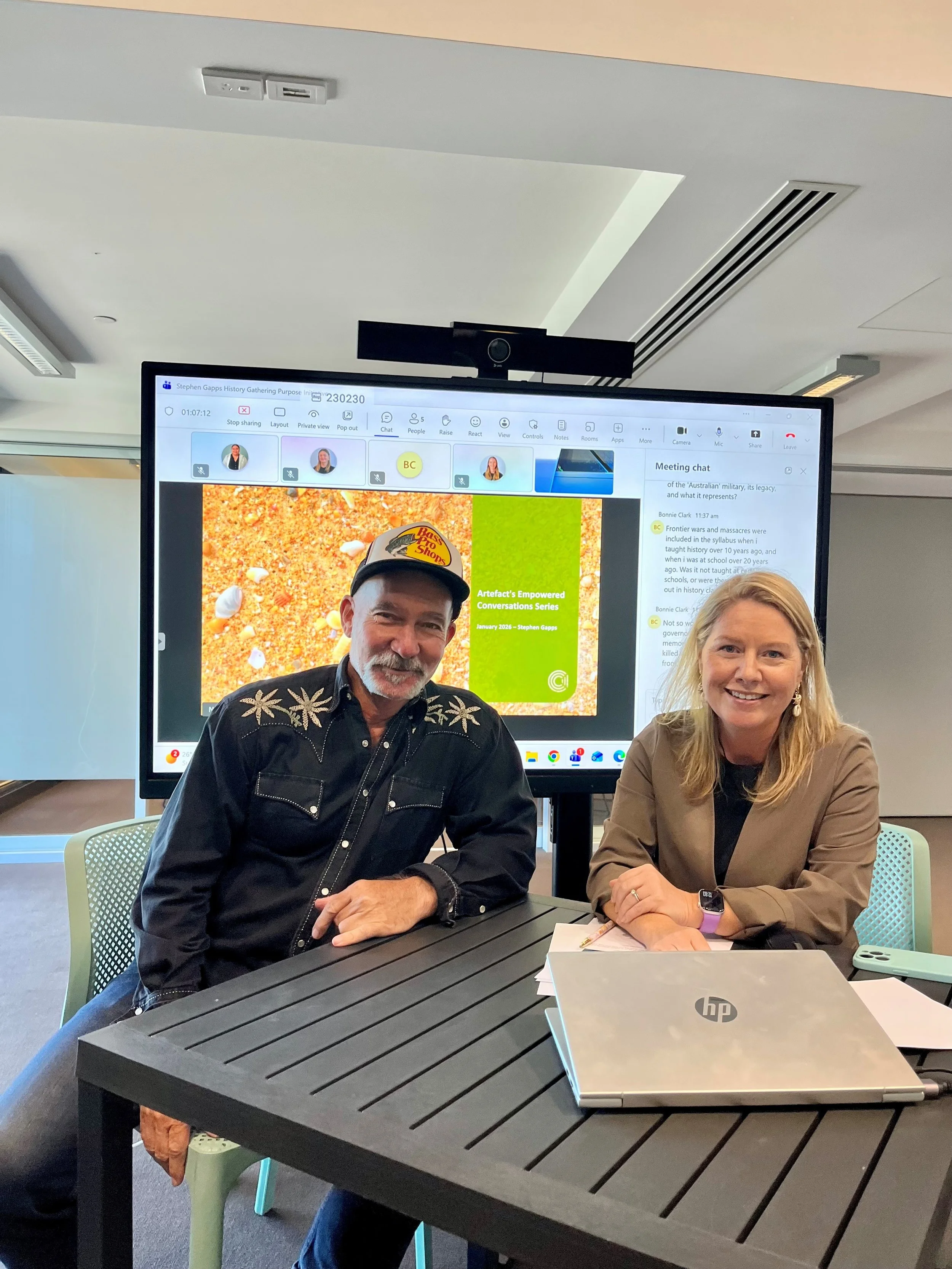 Two people sitting at a black table in a conference room, with a large screen behind them displaying a video call. The person on the left is an older man with a beard, wearing a black shirt with snowflake embroidery and a cap. The person on the right is a woman with blonde hair, wearing a beige jacket and a smartwatch. There is a silver HP laptop on the table.