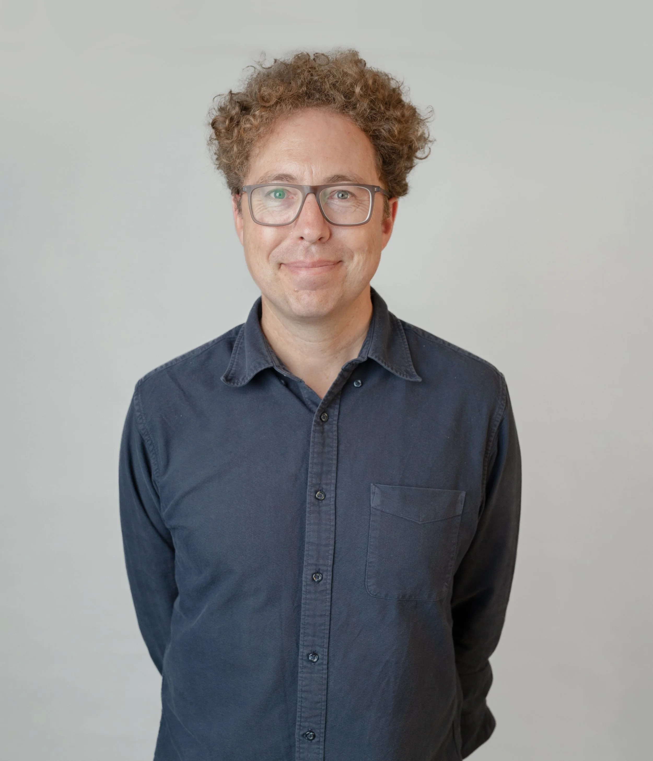 A man with curly brown hair, wearing glasses with clear frames, and a navy blue button-up shirt, standing against a light gray background.