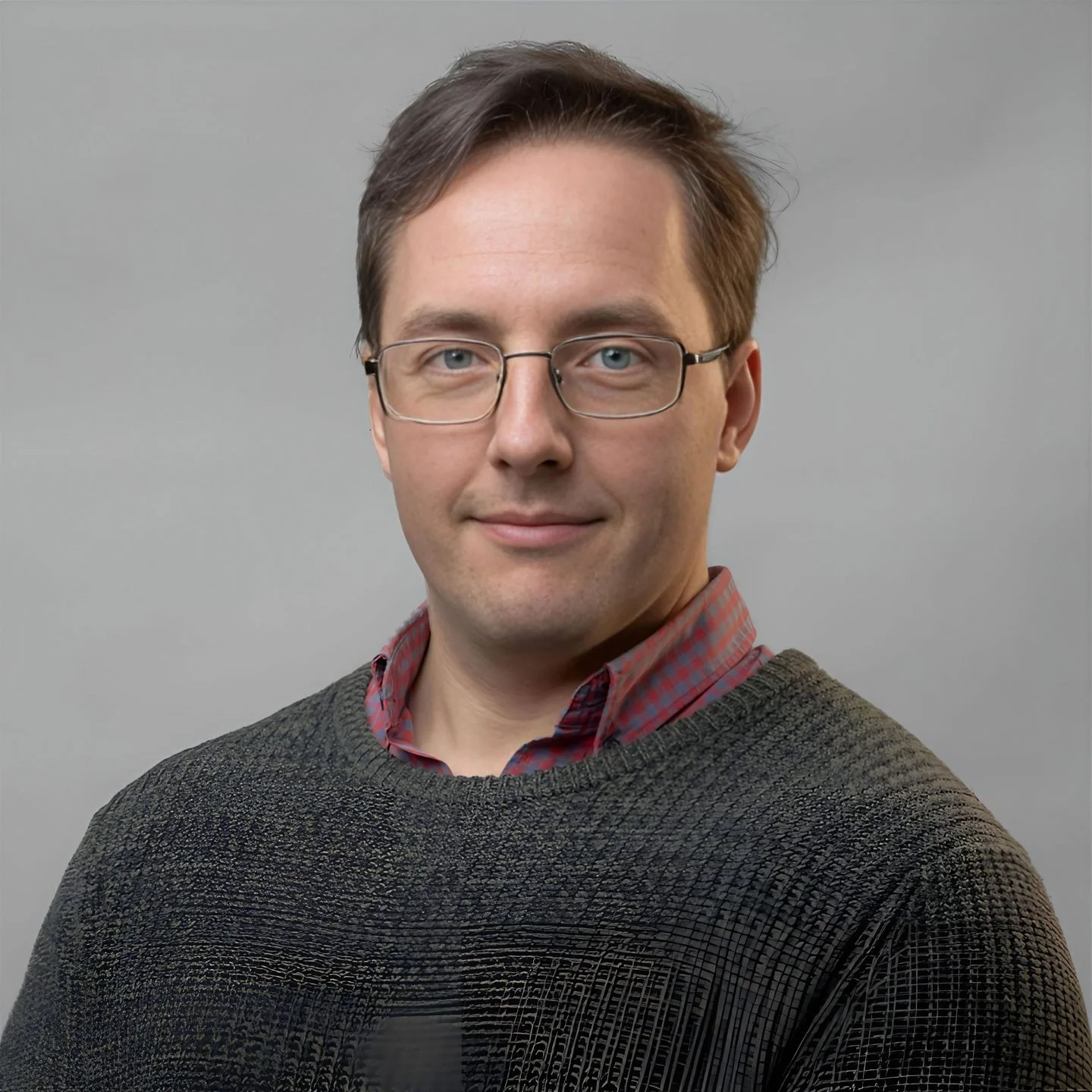 A man with glasses and brown hair wearing a gray sweater and red plaid shirt against a plain gray background.