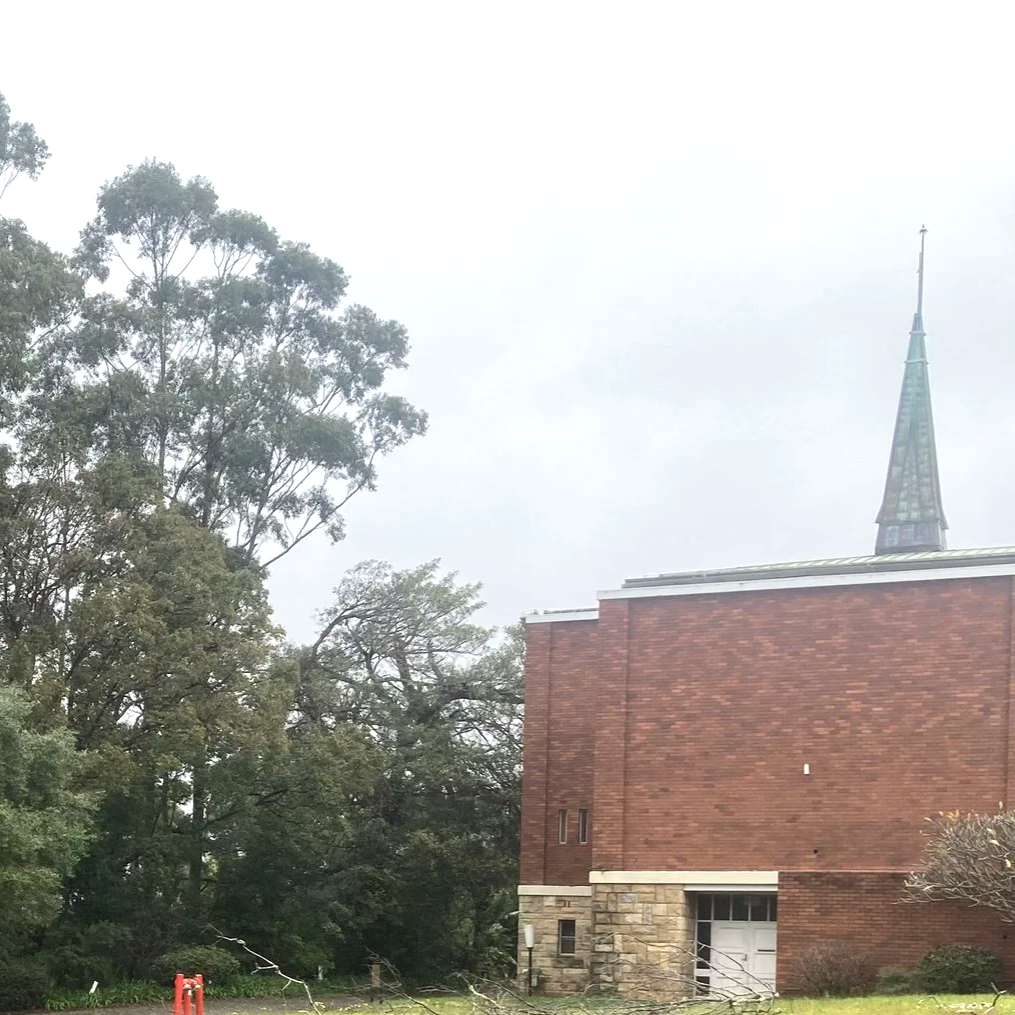 A brick church with a tall steeple, surrounded by trees and cloudy sky.