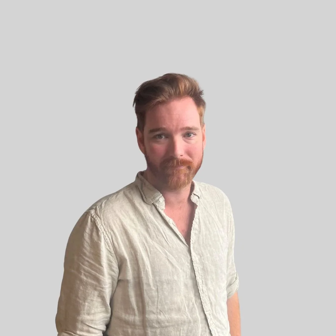 Portrait of a man with red hair and beard, wearing a light-colored shirt, standing against a plain gray background.