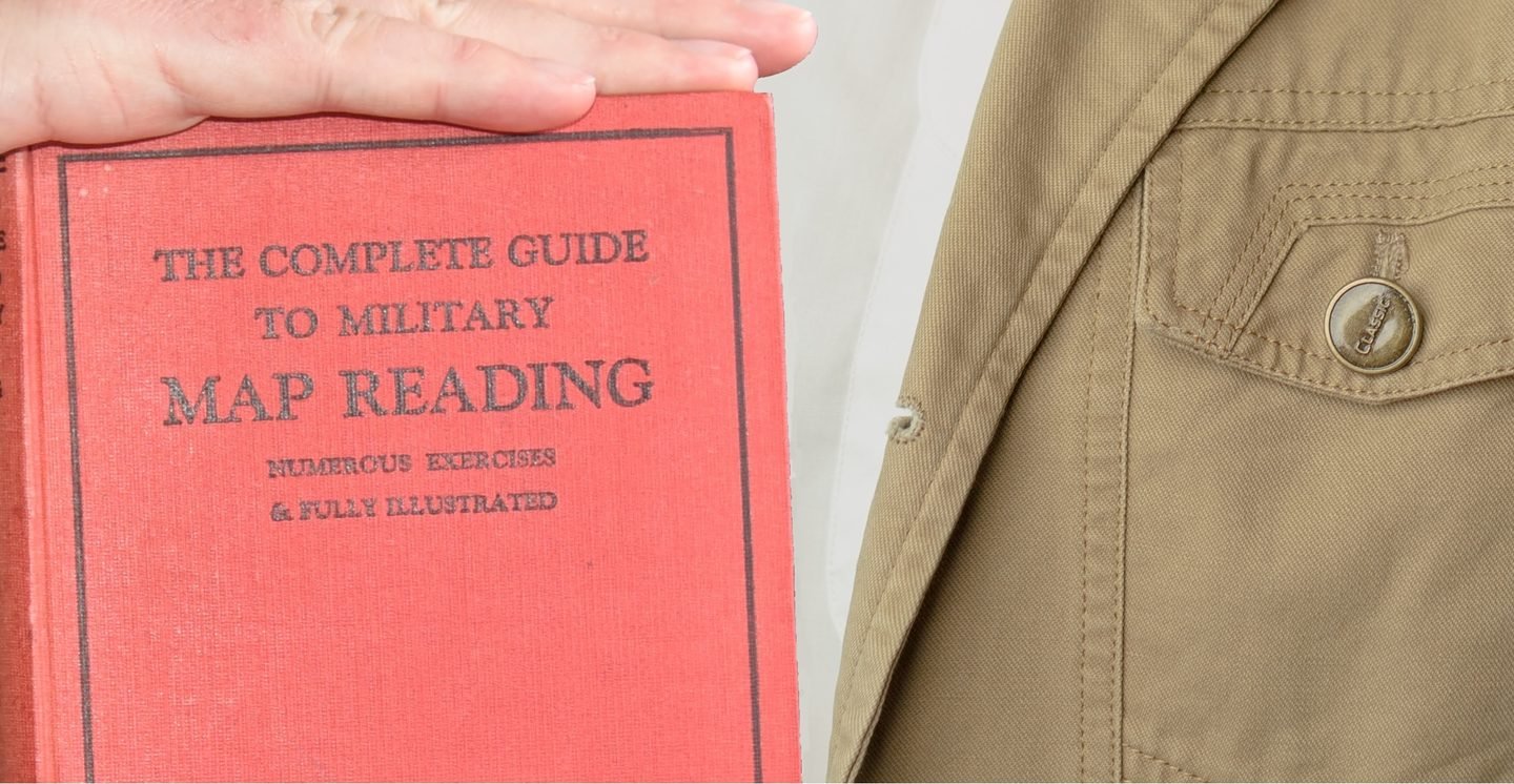 A person holding a red book titled 'The Complete Guide to Military Map Reading' with a beige jacket visible on the right side of the image.