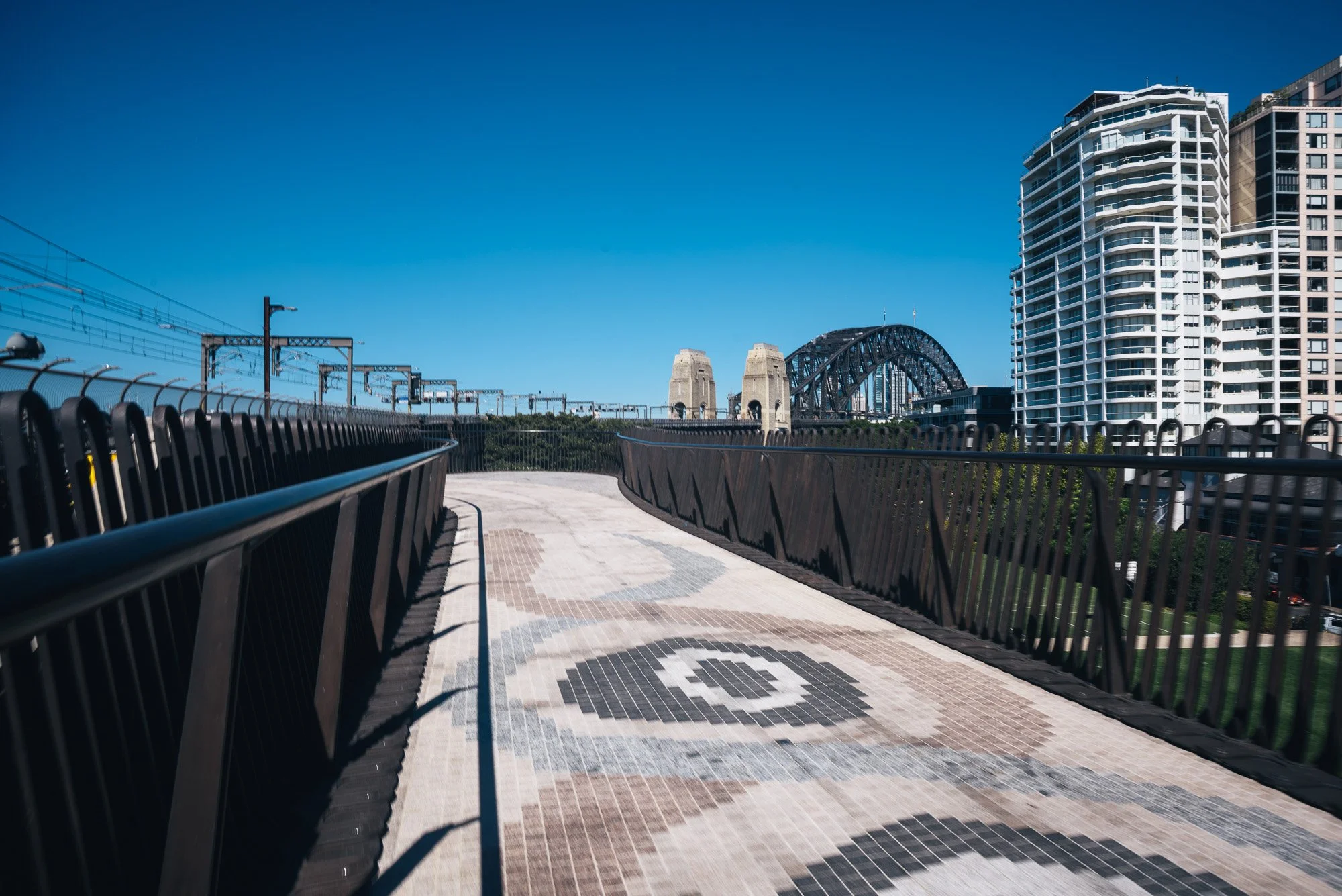 A cityscape viewed from a pedestrian walkway with a patterned ground and black metal railings, with modern high-rise buildings and the Sydney Harbour Bridge in the background under a clear blue sky.
