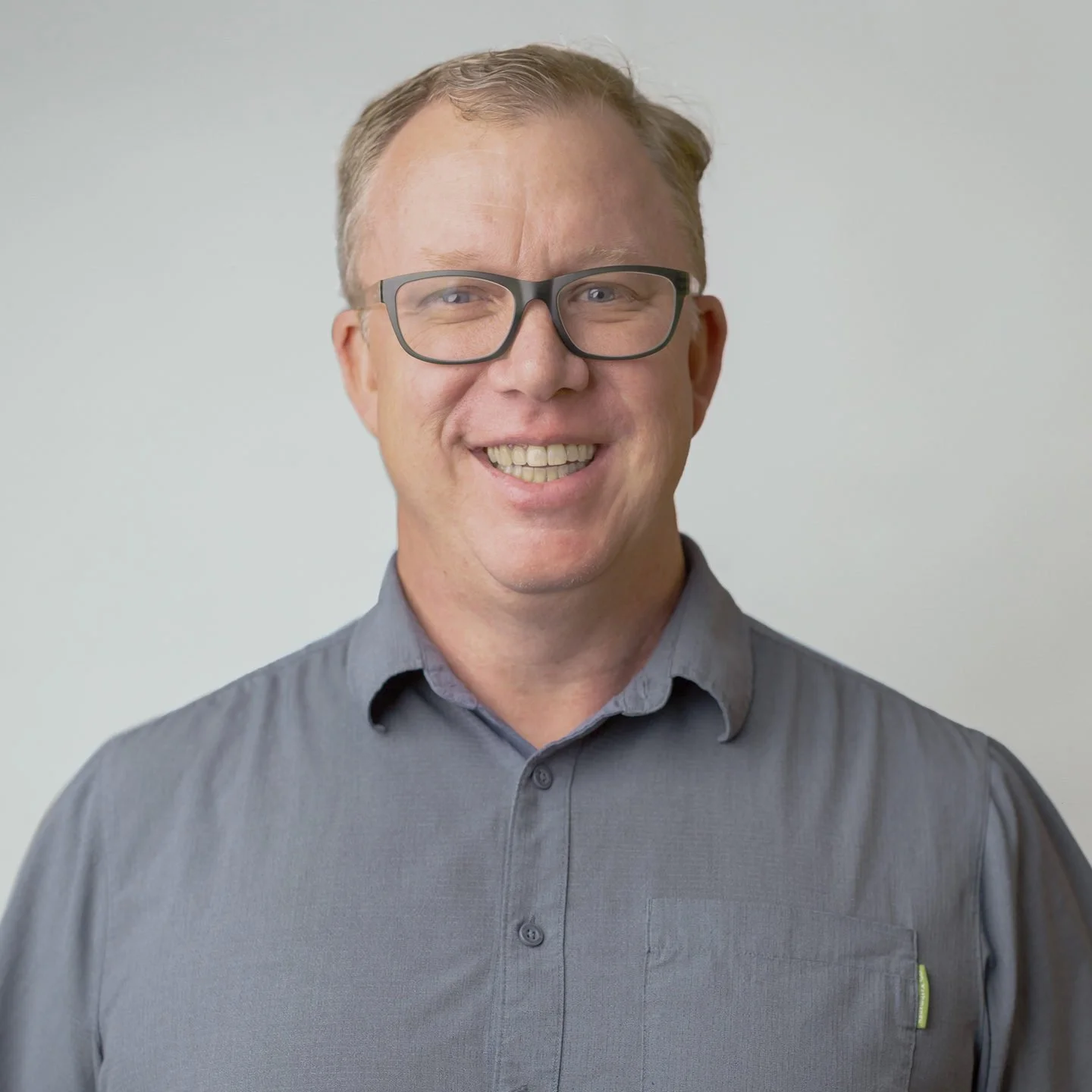 A smiling man with short blond hair, black glasses, and a gray button-up shirt standing against a plain white background.
