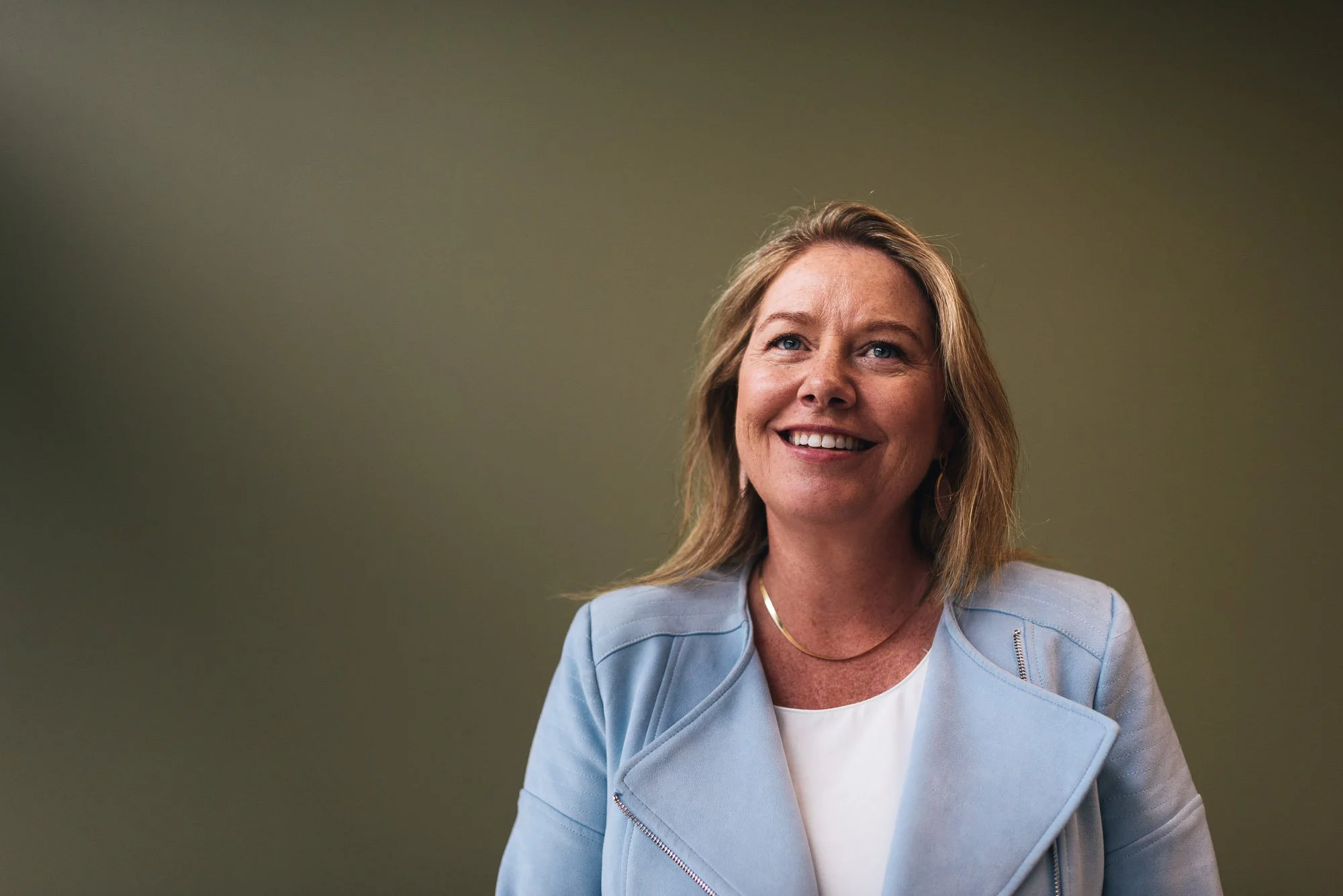 A woman with long blonde hair smiling, wearing a light blue blazer over a white top, standing against a plain greenish background.