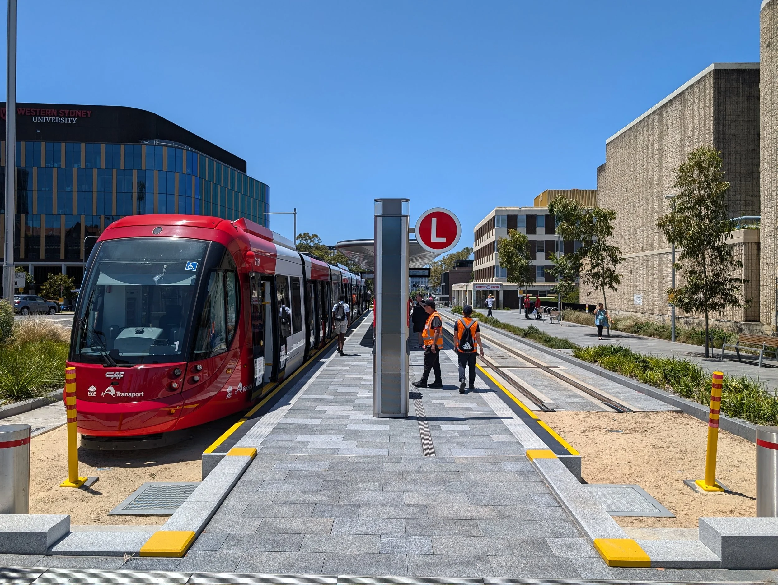 A modern tram station with a red tram, several pedestrians, and a sign indicating a learner driver area, with university buildings and trees in the background under a clear blue sky.
