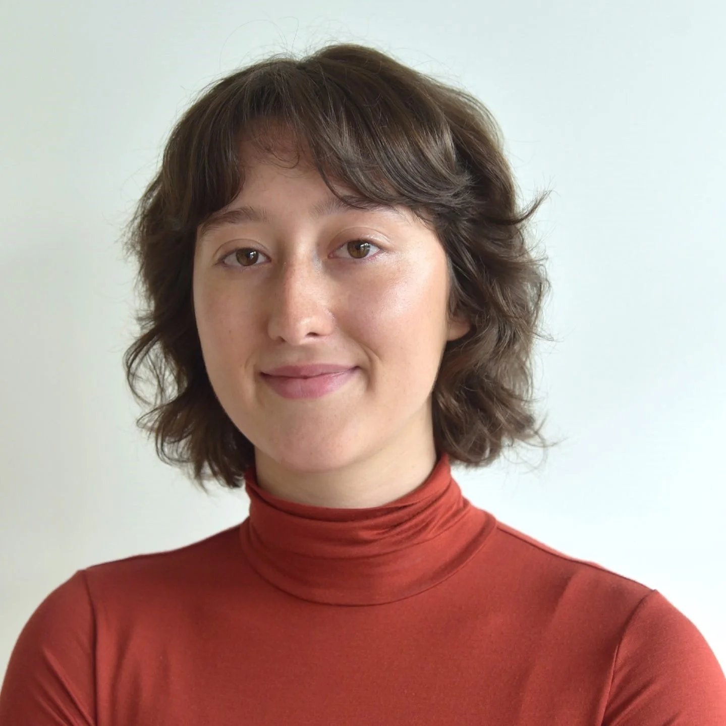 A woman with short, wavy brown hair and brown eyes, wearing a rust-colored turtleneck, smiling softly against a neutral background.