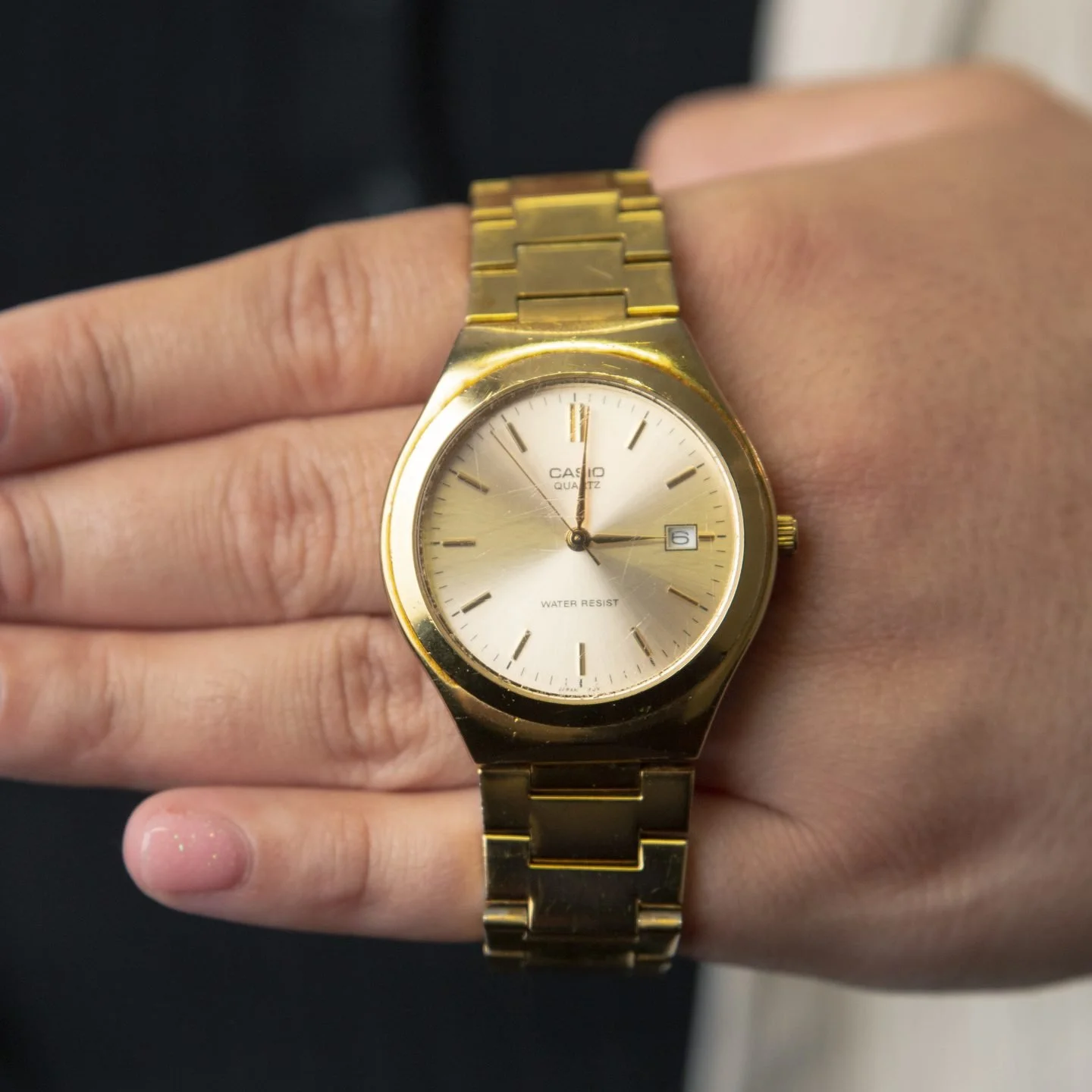 A gold Casio wristwatch on a person's wrist, showing the time as 11:59, with a date window displaying the 6th day, and reading water resistant.