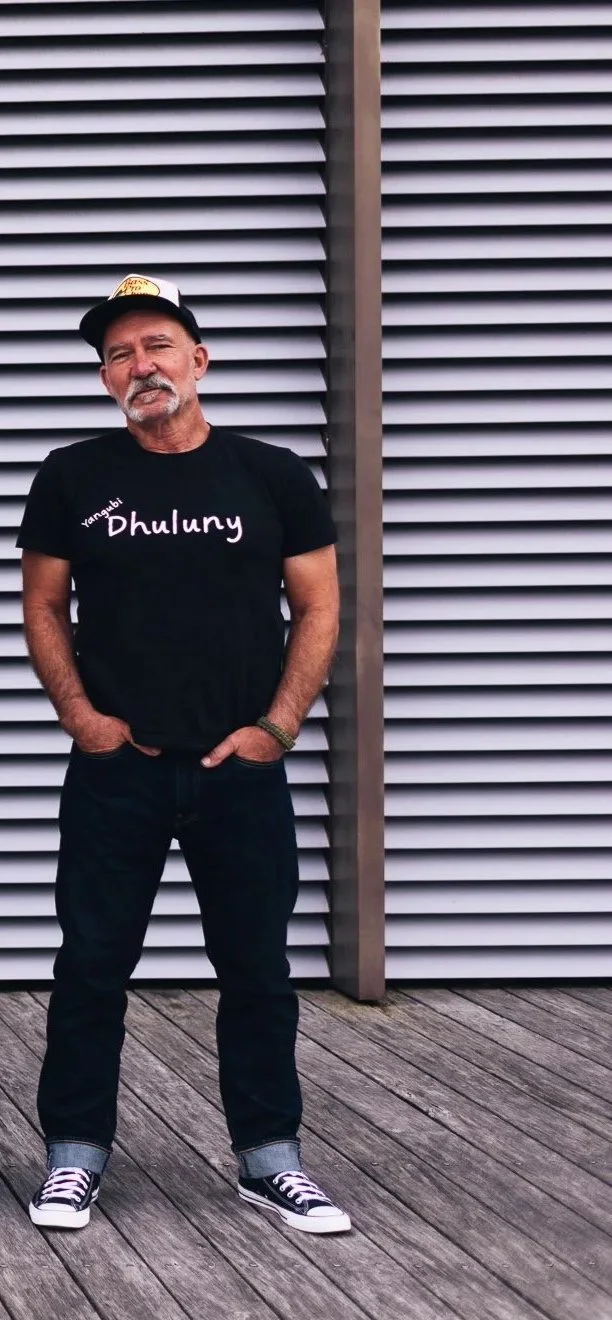 A man with a gray beard and mustache, wearing a black t-shirt, black jeans, black and white sneakers, a baseball cap, and a green bracelet, standing with hands in pockets against a modern metallic wall with horizontal slats on a wooden deck.