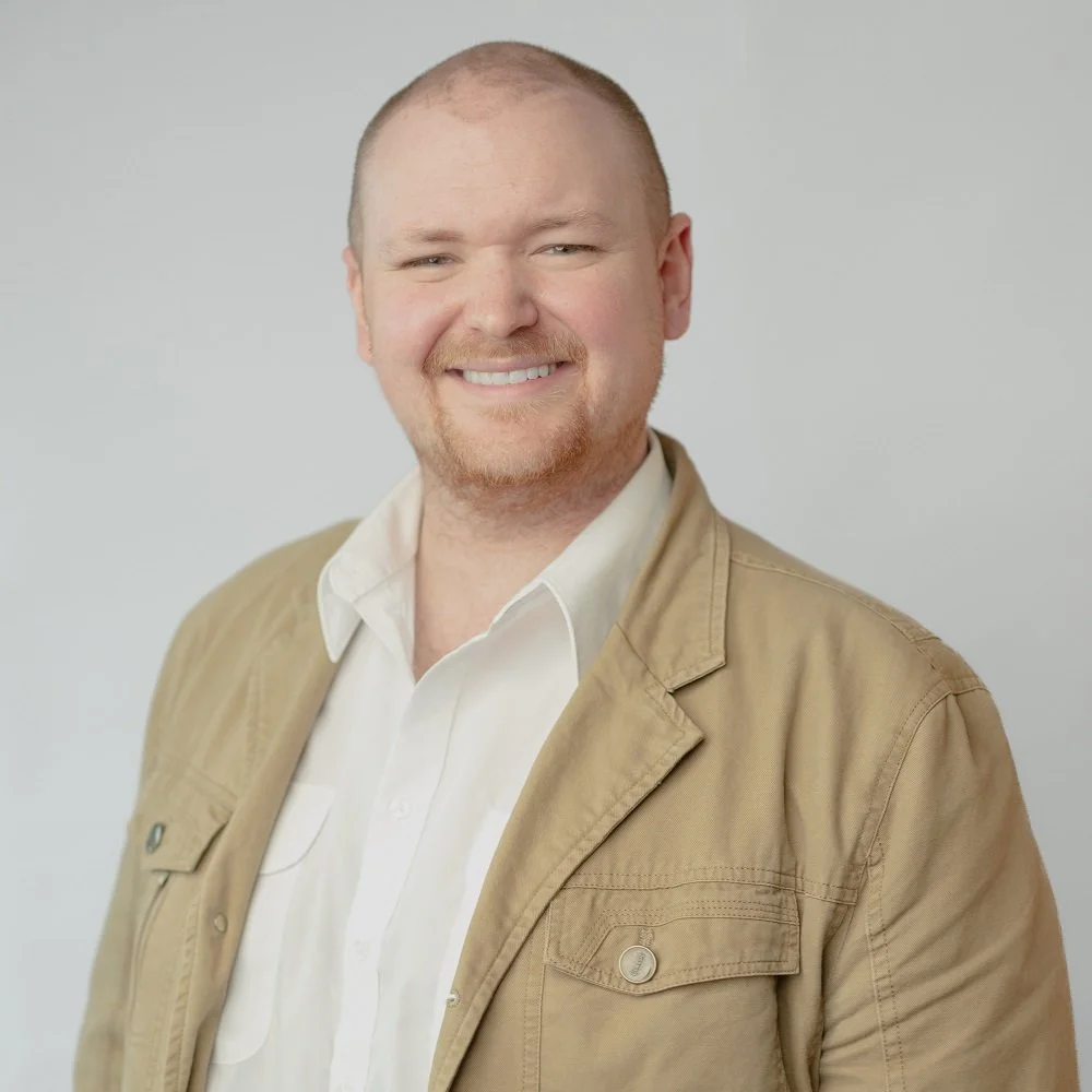 A smiling man with a bald head, reddish beard, and mustache, wearing a beige jacket over a white collared shirt, standing against a plain light gray background.