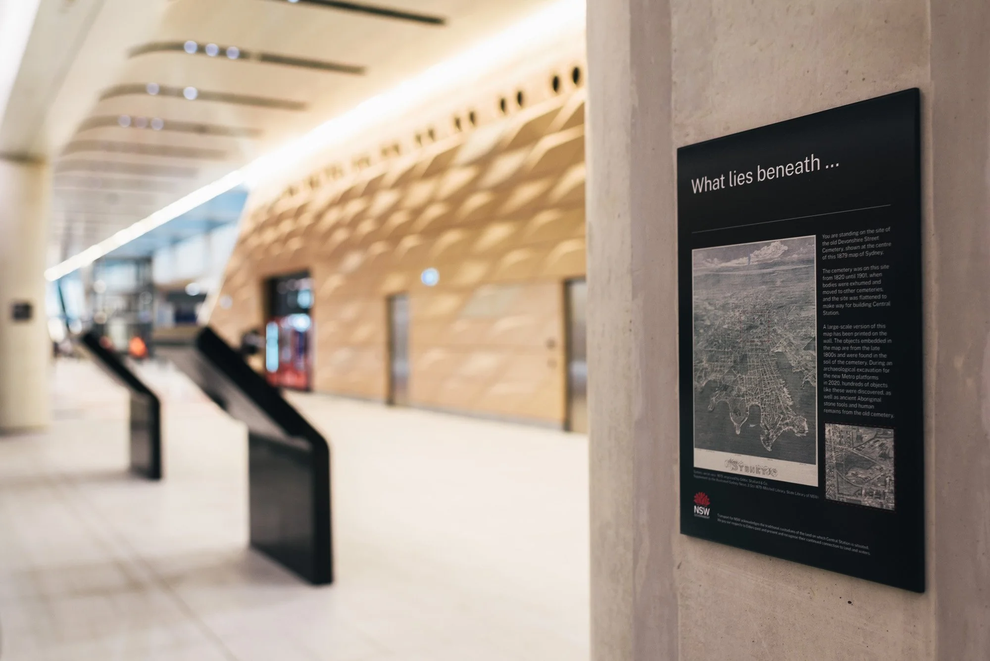 Information signboard titled 'What lies beneath' in a modern indoor setting with wooden architectural features and escalators in the background.