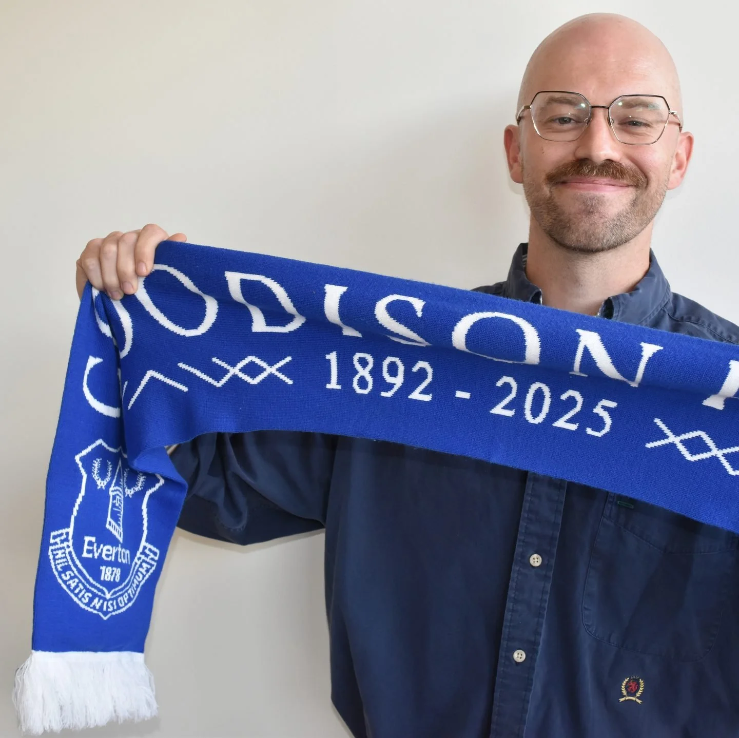 Bald man with glasses smiling, holding an Everton football club scarf with the years 1892-2025, standing in front of a white wall.