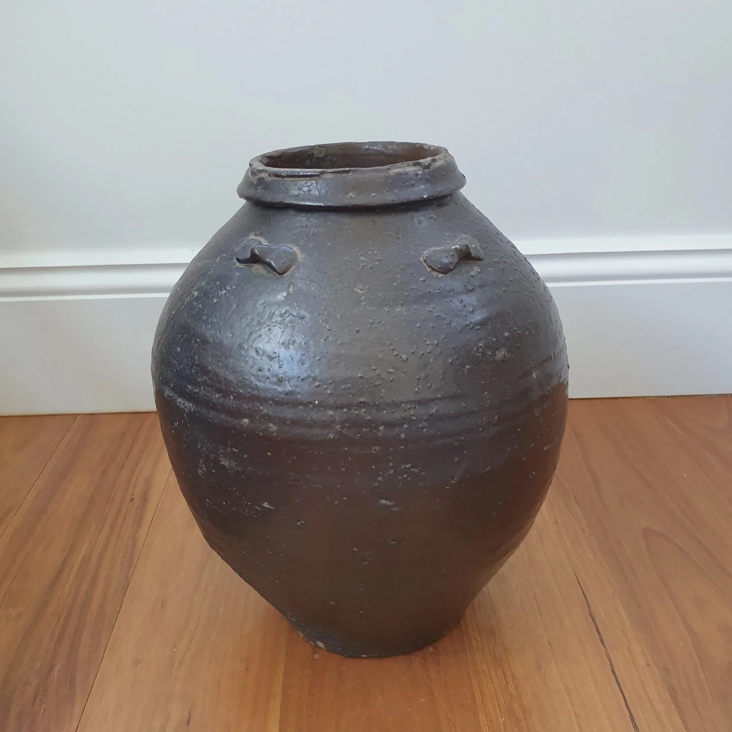 A large, dark brown ceramic storage jar with a rounded body, narrow top, and small protruding handles on either side, sitting on a wooden floor against a white wall with baseboard.