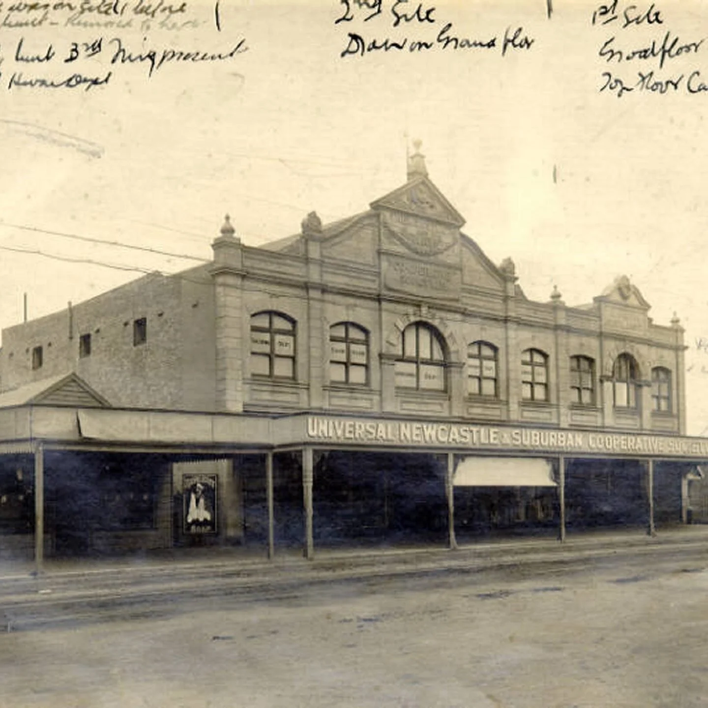 An old sepia-toned photograph of a two-story building labeled 'Universal Newcastle Suburban Cooperative Society'. The building has arched windows and decorative architectural details on the front, with a covered sidewalk in front supported by posts. There is a small poster or sign on the sidewalk and handwritten text at the top of the photo.