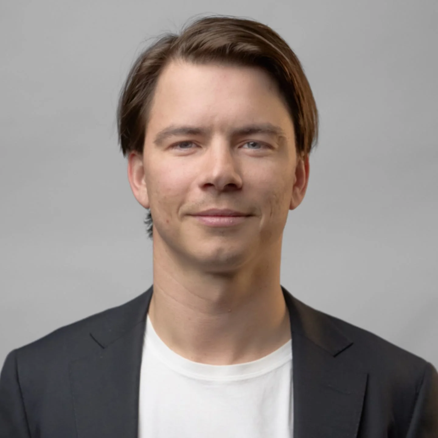 A portrait of a young man with brown hair, wearing a black blazer over a white t-shirt, against a plain gray background.