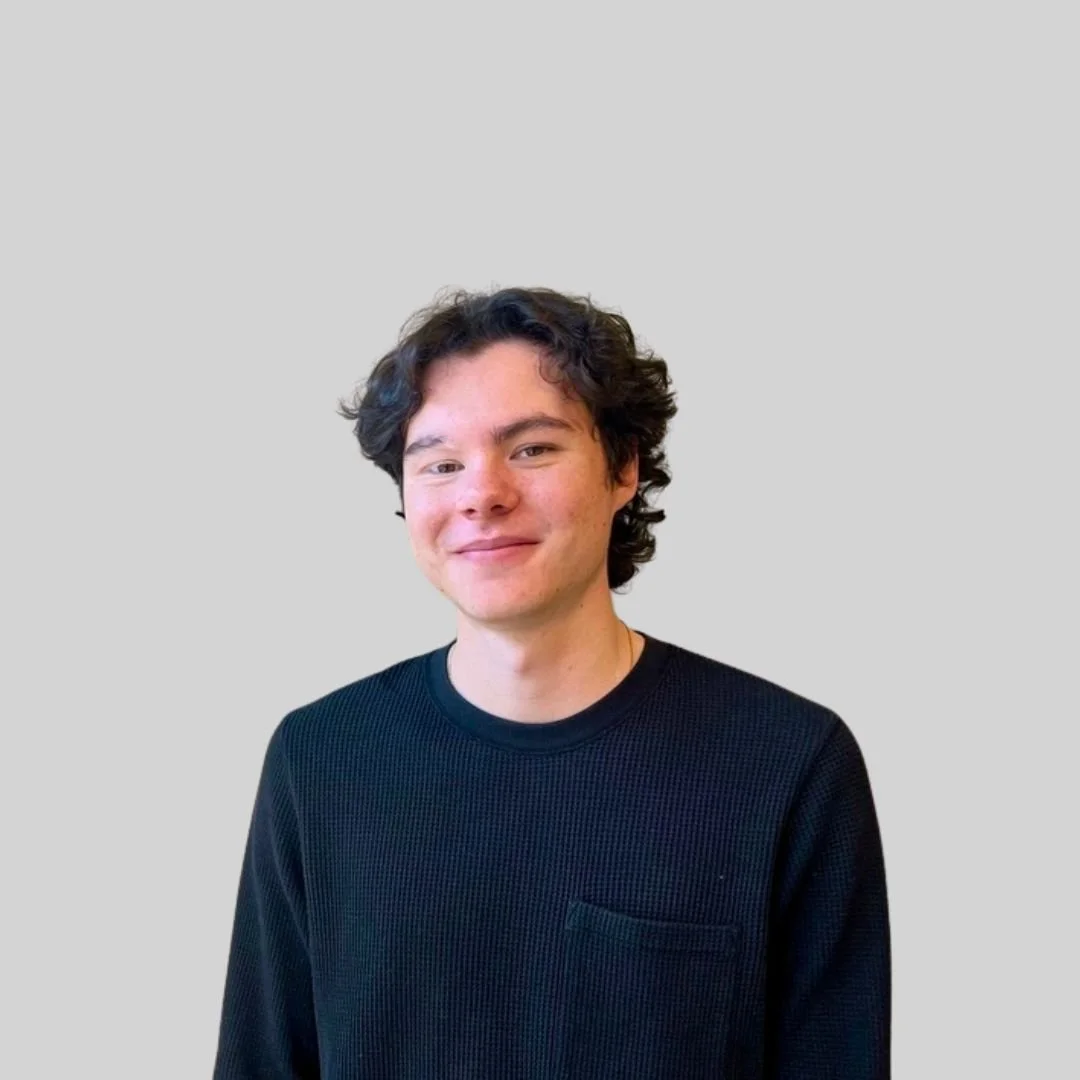 A young man with curly dark hair, wearing a black sweater, smiling, standing against a plain light gray background.