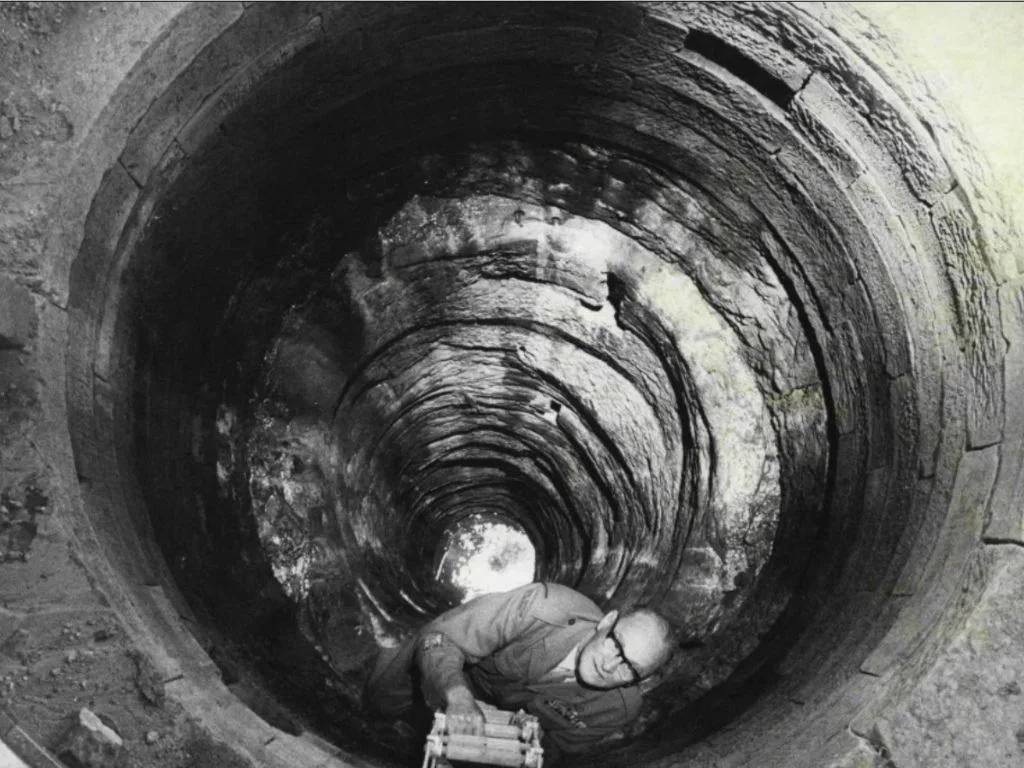 A worker wearing protective glasses and a jacket crouches inside a deep, circular stone well or tunnel, with the circular pattern of the stone lining visible all around.