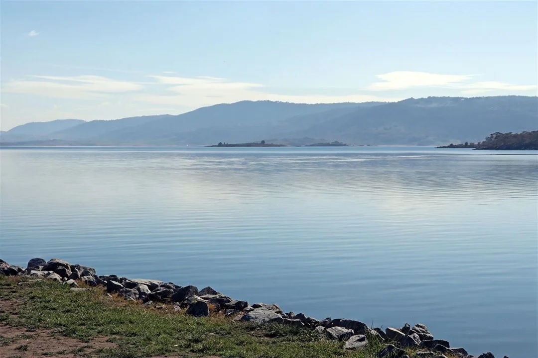 A calm body of water with gentle ripples, bordered by a rocky shoreline and grassy land in the foreground. In the background, there are distant mountains under a partly cloudy sky.