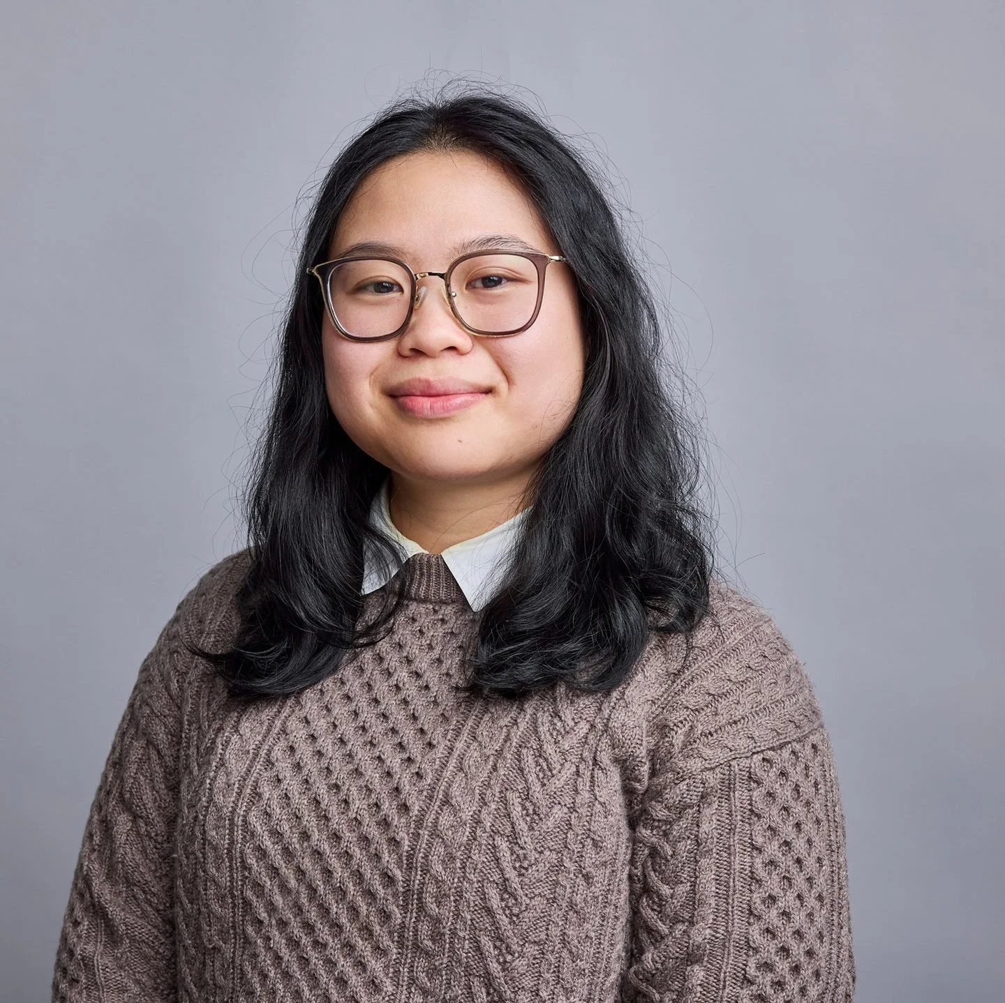 A woman with black wavy hair, wearing glasses, a white collared shirt, and a brown knitted sweater, smiling at the camera against a plain gray background.