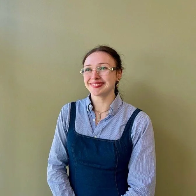 A young woman with glasses smiling, wearing a light blue button-up shirt under a dark blue overall dress, standing against a plain beige wall.