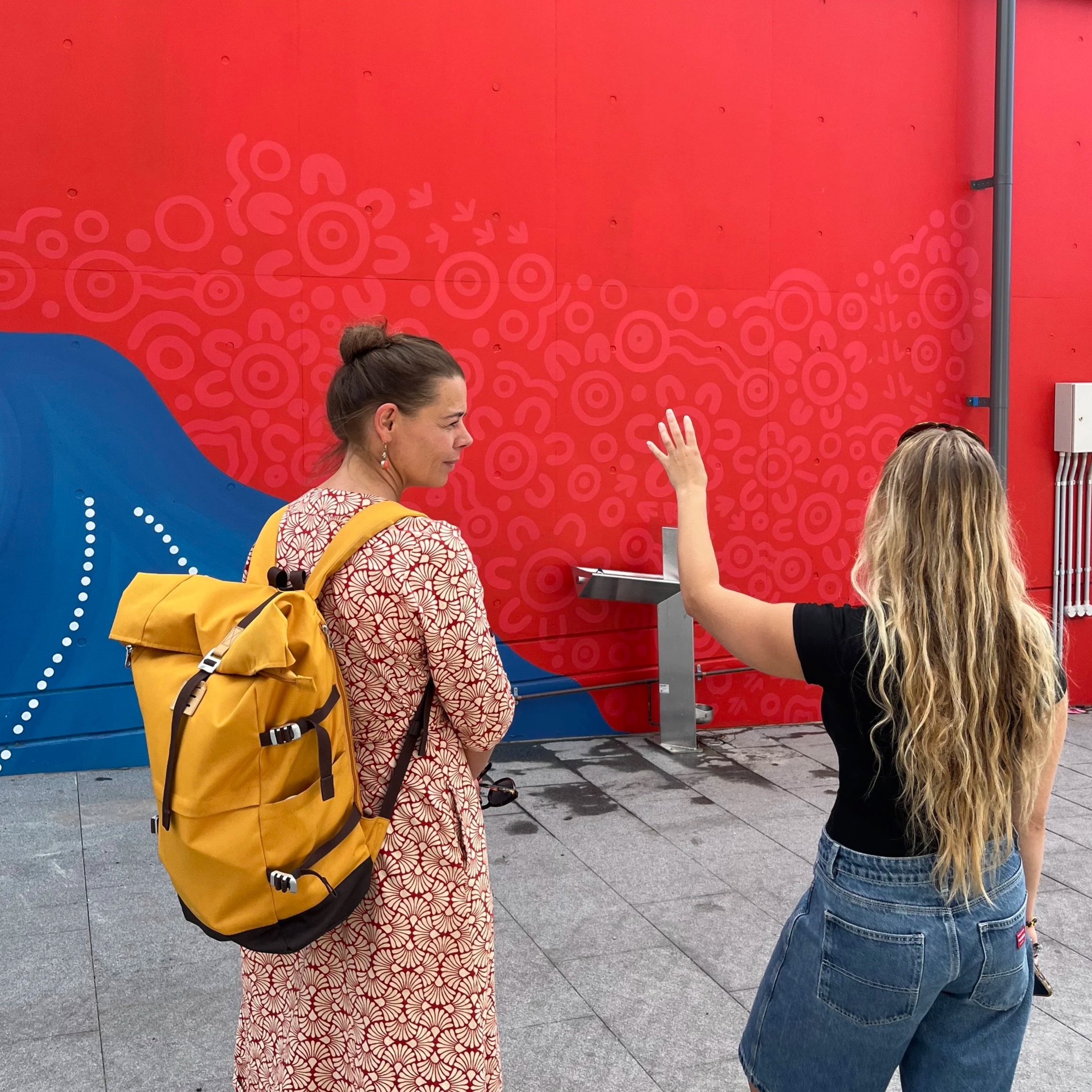 Three women standing outdoors with a red wall background, two with long hair, one with hair tied up, one woman wearing a yellow backpack.