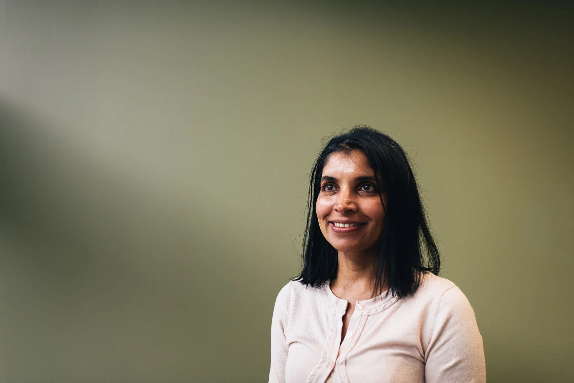 A woman with long dark hair, smiling, wearing a light pink blouse, standing against a plain beige wall.