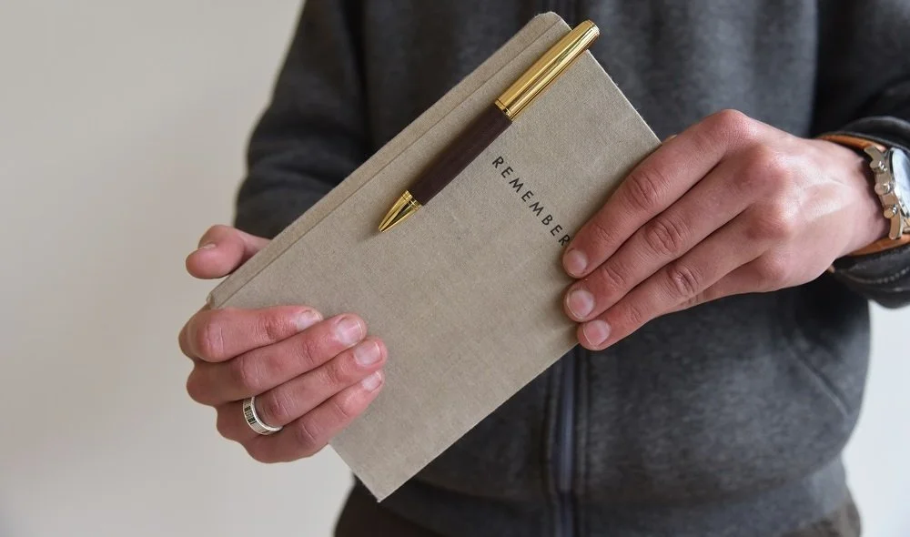 Person holding a beige notebook with a gold and dark brown pen clipped onto it, and the word 'REMEMBER' printed on the cover.