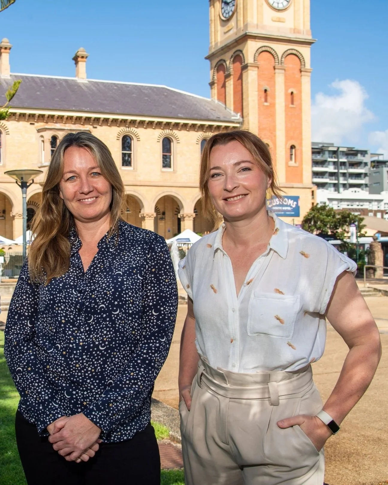 Two women standing outdoors in front of a historic building with a clock tower. The woman on the left has long blonde hair and is wearing a dark blue shirt with a star and moon pattern. The woman on the right has reddish hair tied back and is wearing a white shirt with small print and beige pants.