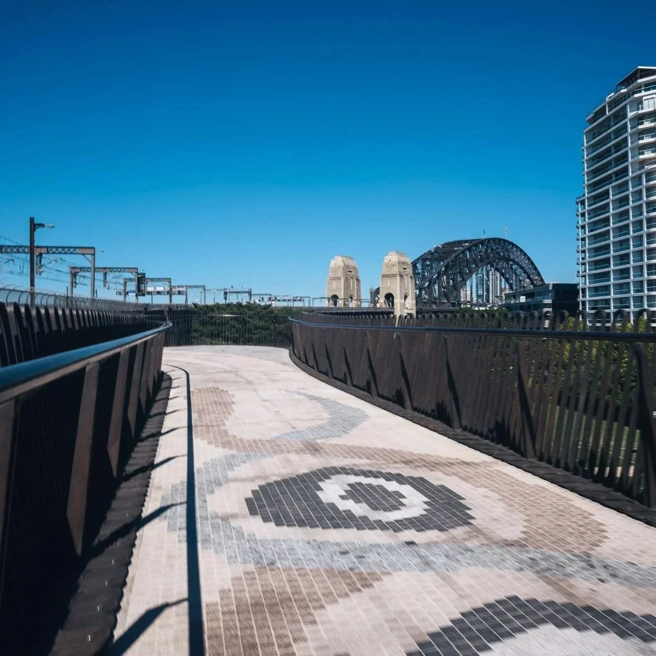 Empty walkway with geometric tile design, black metal railing, Sydney Harbour Bridge visible in the background, surrounded by modern high-rise buildings against a clear blue sky.