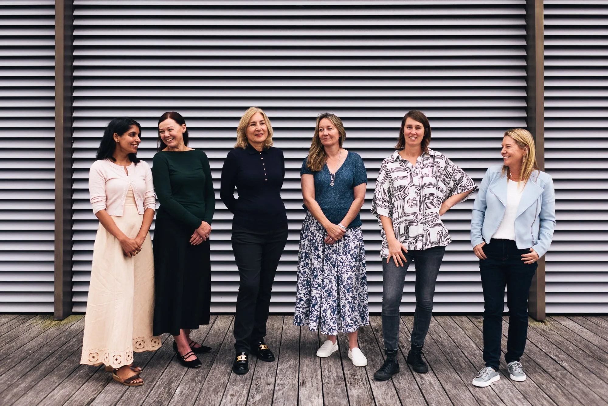 Group of six women standing in front of a modern, horizontally slatted wall. They are dressed in a mix of casual and semi-formal clothing, and are smiling or looking at each other.