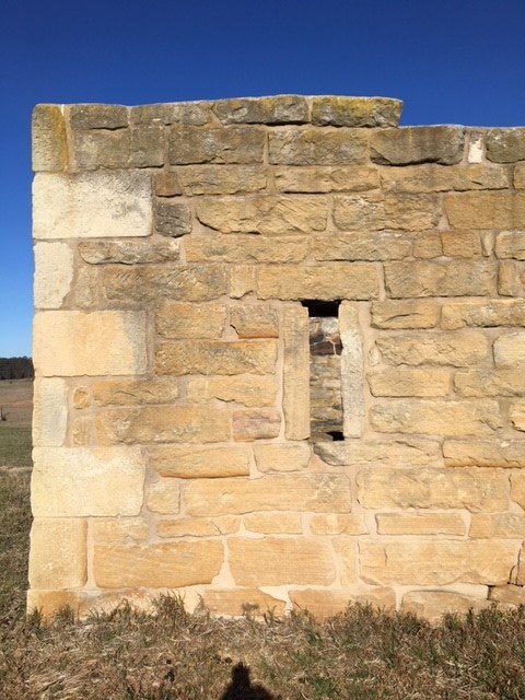 A section of a beige stone wall with a rectangular window opening, set against a bright blue sky and dry grass ground.