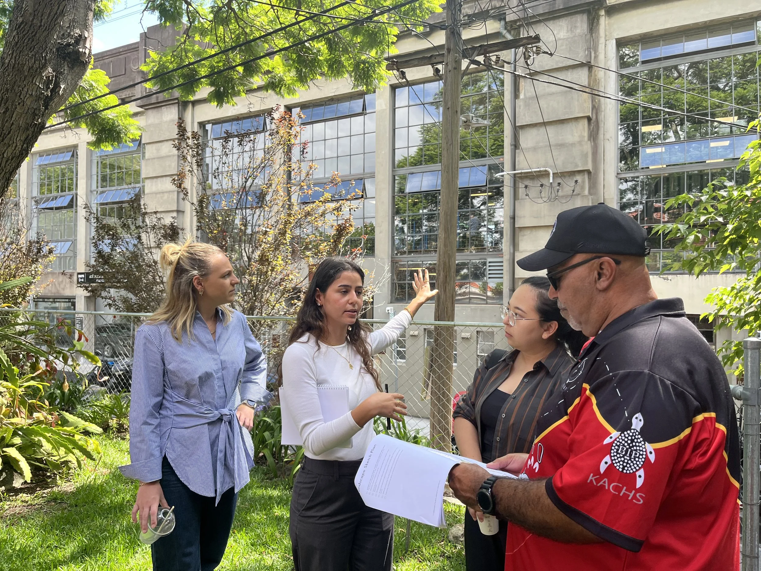 A group of people having a discussion outdoors in a park with greenery and a building with large windows in the background. One person is holding papers, and others are engaging and listening.