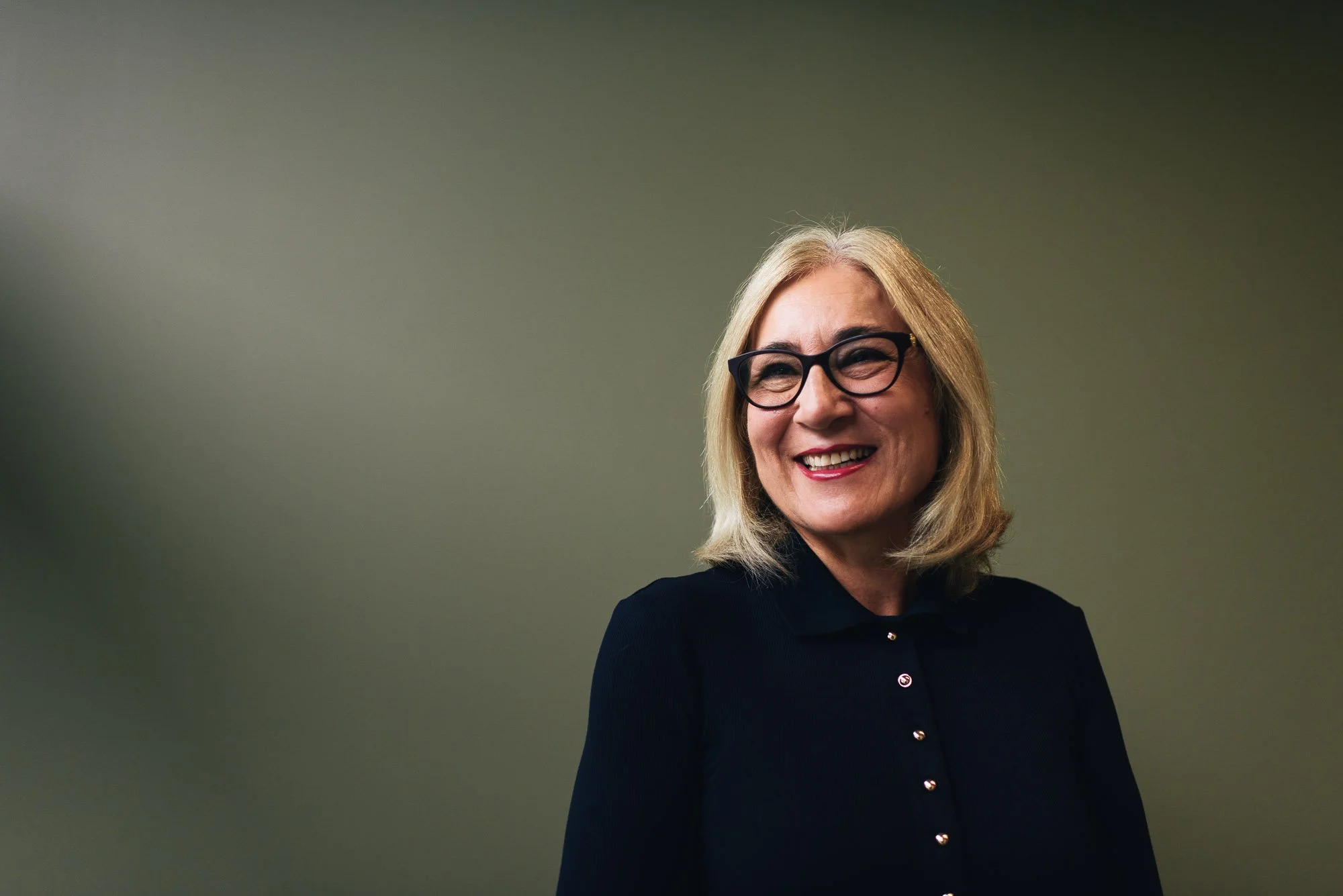 Artefact Industry Engagement Director Anita Yousif,  smiling with blonde hair, glasses, and a black jacket standing against a plain, gradient background.