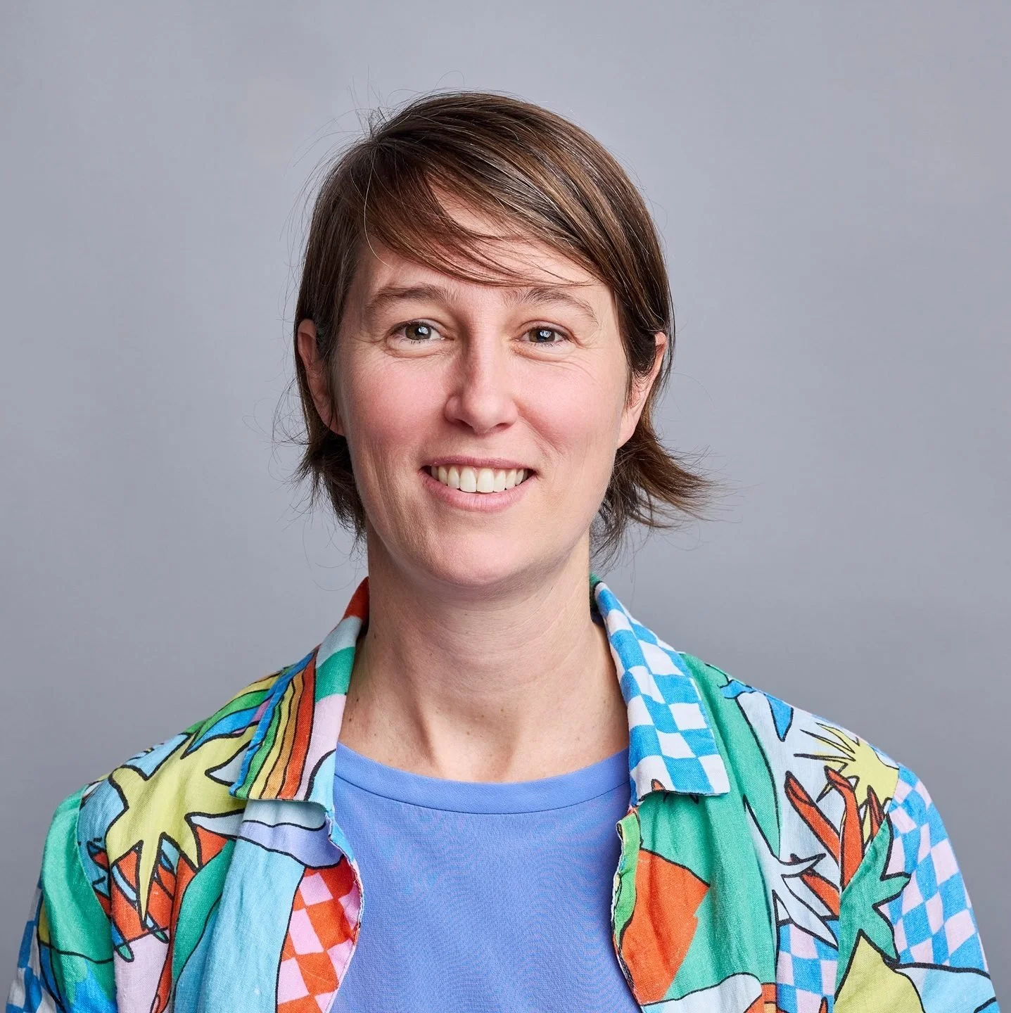 A woman with short brown hair smiling, wearing a colorful, patterned shirt over a blue T-shirt, against a plain gray background.