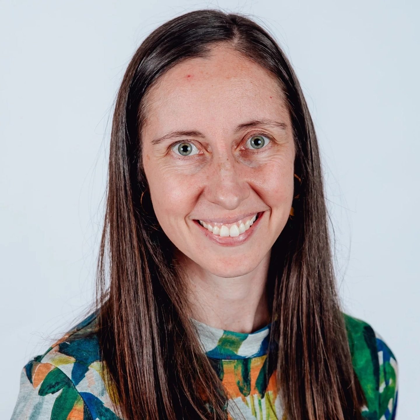 Close-up portrait of a smiling woman with long brown hair, blue eyes, wearing a colorful patterned shirt against a plain white background.
