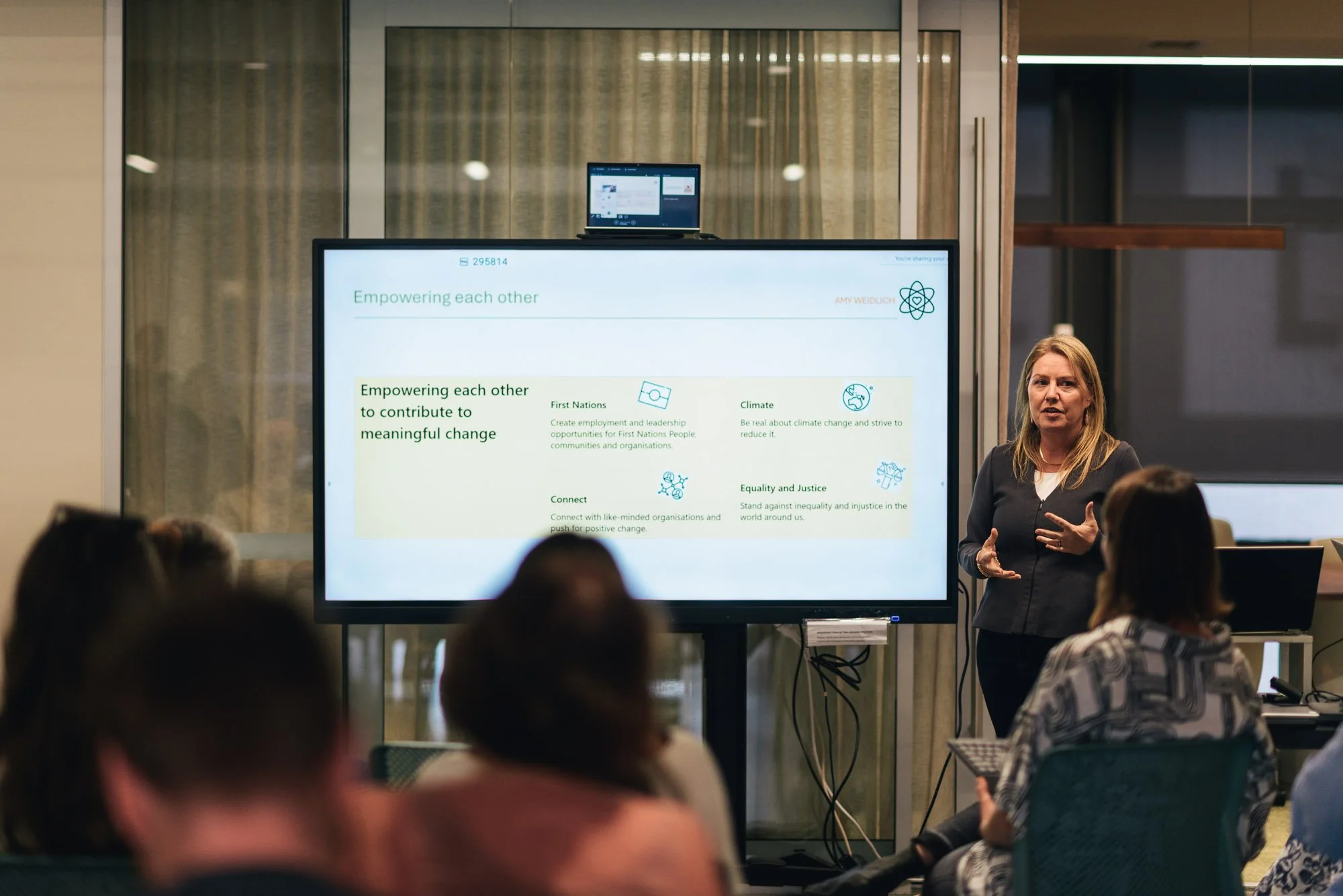 A woman giving a presentation in a conference room with a large screen displaying a slide titled "Empowering each other". Audience members are seated, facing the speaker and screen.