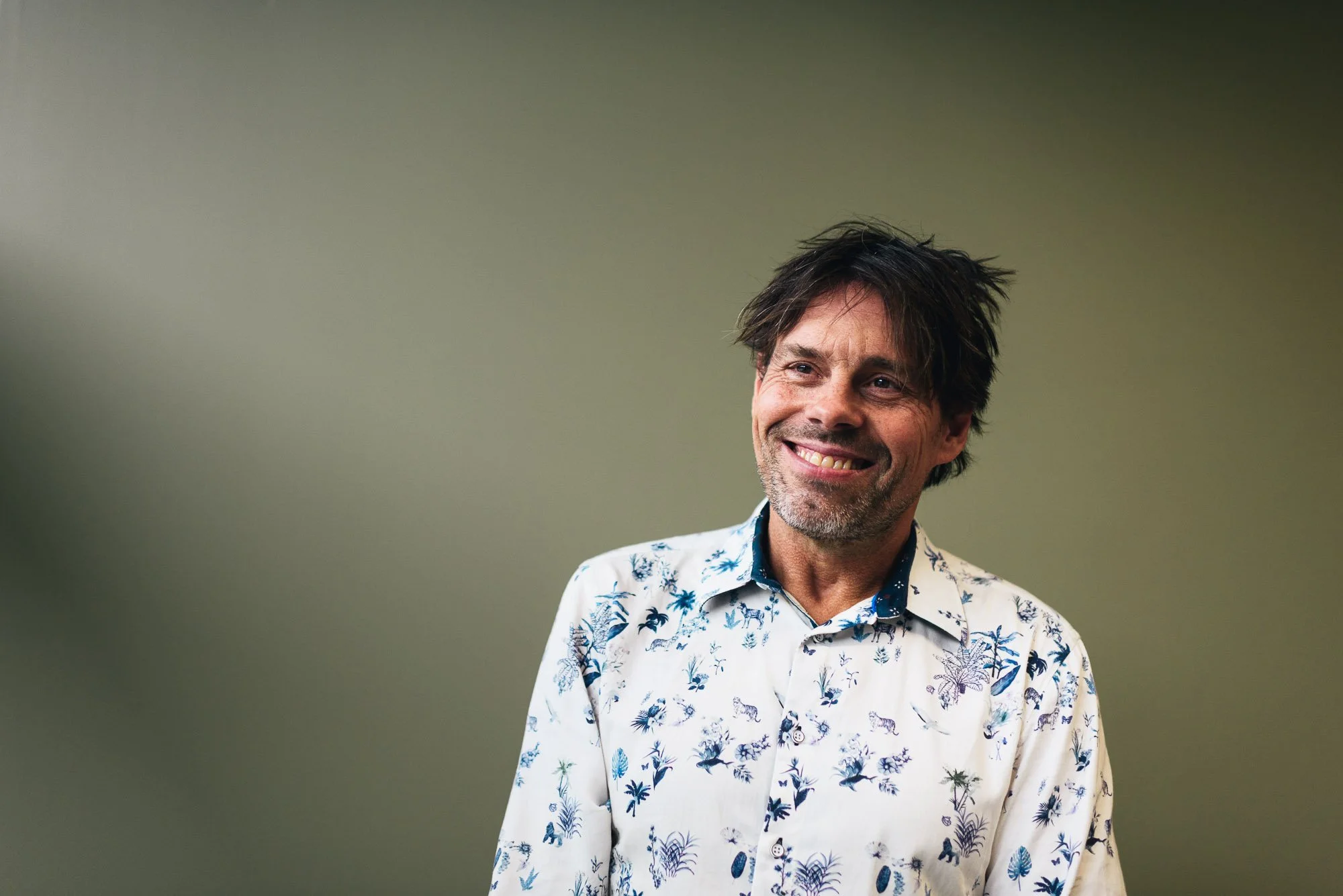 Smiling man with dark hair and stubble wearing a white shirt with blue and purple floral patterns, standing against a plain light gray-green background.